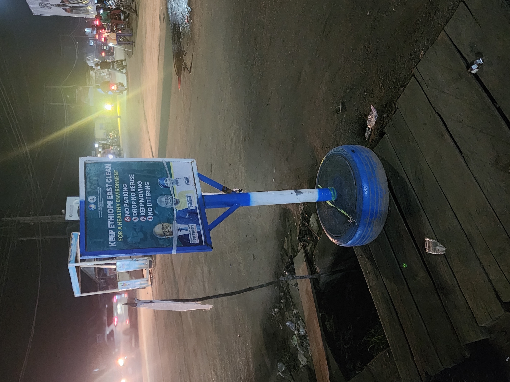 At night in Obiaruku, Nigeria, a sign promoting cleanliness in Ethiope East stands near a roadside drain. The sign, partially obscured by the angle, displays the message "KEEP ETHIOPE EAST CLEAN FOR A HEALTHY ENVIRONMENT" along with instructions prohibiting parking, dropping refuse, and littering. Images of individuals named Oborevwori, Ogedegre, and Charles are also visible on the sign. The sign is attached to a pole supported by a blue-painted tire acting as a base. The immediate environment consists of a paved road and wooden planks covering a drain. Debris and litter are scattered around the drain. In the background, streetlights illuminate a row of parked motorcycles and other vehicles, indicating a moderately busy street scene.