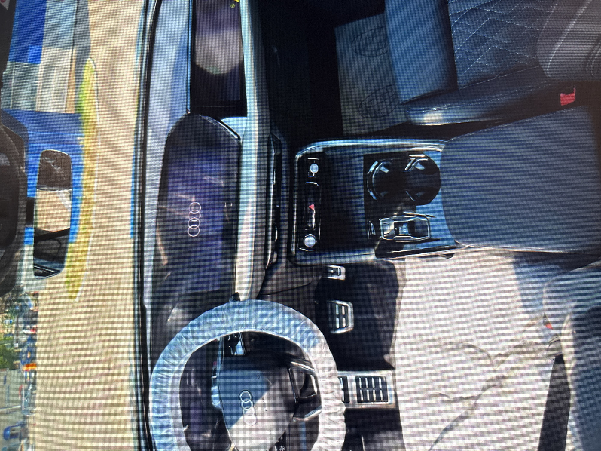 The image shows the interior of a new car, likely an Audi, parked outdoors on a sunny day. The steering wheel is covered with a protective white cloth, and the dashboard features a large digital display showing the Audi logo. The center console has a gear shifter, cup holders, and climate controls. The seats are upholstered in dark leather with a diamond-stitched pattern. The car appears to be in a parking lot with a paved surface and some vegetation in the background. There are no people visible in the image. The location is identified as Moscow, Russia.