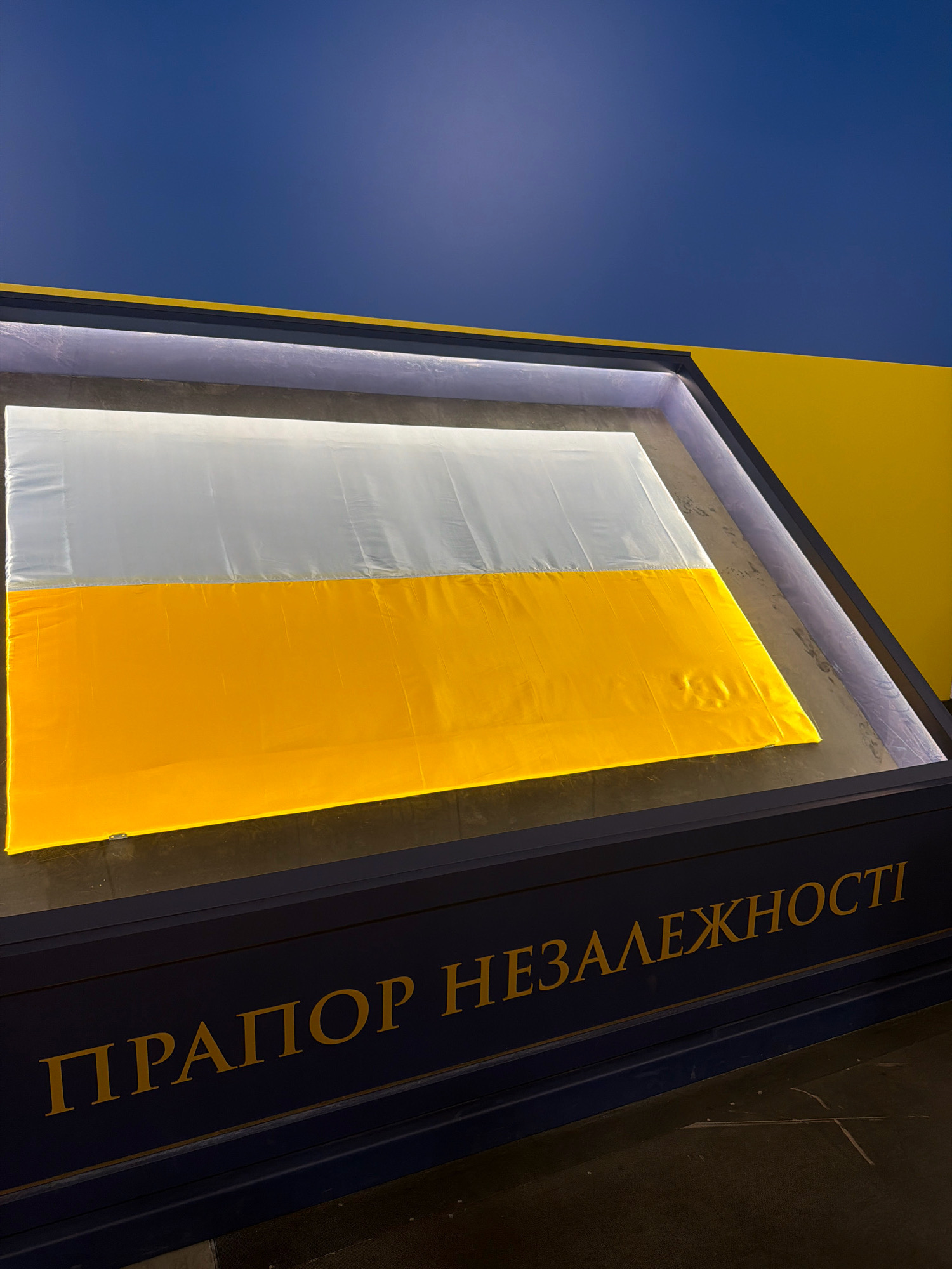 An image shows a flag displayed in a recessed area with a yellow and blue color scheme. The flag is horizontally divided into two equal halves: the top half is white and the bottom half is yellow. The flag is illuminated from above, casting a bright glow. The surrounding display area is framed by a dark blue border with yellow text that reads "ПРАПОР НЕЗАЛЕЖНОСТІ", which translates to "FLAG OF INDEPENDENCE". The background consists of a dark blue wall and a yellow wall on the right. The image is taken from a slightly elevated perspective, looking down at the flag. The overall impression is that of a museum exhibit or a public display celebrating a national symbol. The location of this display in Kyiv, Ukraine, is not specified within the image itself, but the text on the display and the flag's colors (white and yellow, often associated with Ukraine) suggest a Ukrainian context.