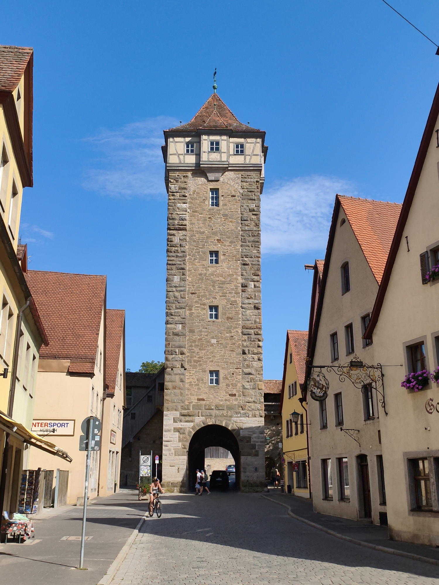The image shows the Plönlein, a prominent landmark and iconic street intersection in the medieval town of Rothenburg ob der Tauber, Germany. The scene is dominated by the Siebers Tower, a tall, stone structure with a red-tiled roof, which forms a striking archway over the cobbled street. To the left, a yellow building with a red-tiled roof features a sign that reads "INTERSPORT HUBSCH". Further down the street, a woman on a bicycle is riding towards the tower, and several other pedestrians are visible walking near the archway. The buildings lining the street are characteristic of medieval German architecture, with their steeply pitched roofs and timber-framed upper stories. The sky is a clear blue with a few scattered clouds, indicating a bright, sunny day. The overall impression is one of historical charm and picturesque beauty.