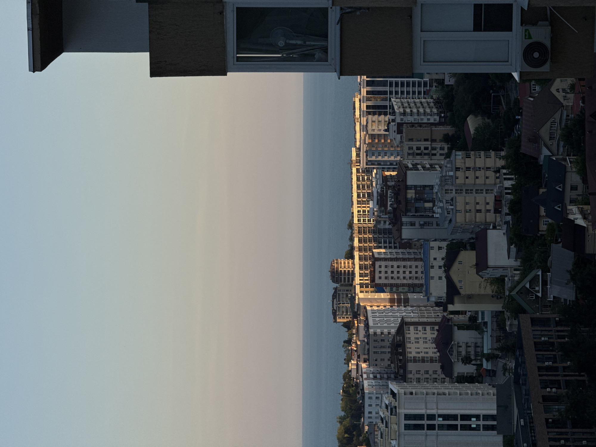 This vertical photograph depicts a coastal cityscape in Sochi, Russia, observed during what appears to be late afternoon or early evening, characterized by a warm, subdued light. The image is horizontally segmented into three main areas: sky, sea, and an urban landscape.
The upper portion features a clear sky, transitioning from a light blue at the zenith to a pale, warm yellow-orange hue near the horizon. Below the sky is a calm, dark blue expanse of the Black Sea, forming a distinct horizon line.
The dominant midground and foreground display a dense urban environment comprised of numerous multi-story buildings, predominantly residential or hotel structures. These buildings exhibit varied architectural styles and colors, ranging from lighter facades on the left to darker, shadowed areas in the lower parts of the frame. Several buildings, particularly those further back and to the right, are illuminated by direct, low-angle sunlight, casting warm golden tones on their upper levels and facades. Some structures appear to be newer constructions or undergoing development. Interspersed between the buildings are pockets of green foliage, indicating urban trees and landscaping.
On the far right side of the image, a partial view of a light brown building facade occupies the foreground. This section of the building is largely in shadow. A rectangular window with a white frame is visible, inside which indistinct shapes, possibly a fan or curtains, are discernible. Directly below this window, a "Bollu" brand "DC Inverter" air conditioning unit is mounted on the exterior wall.
No people, vehicles, or explicit activities are visible within the frame. The scene conveys a quiet, expansive view of a developed coastal city.