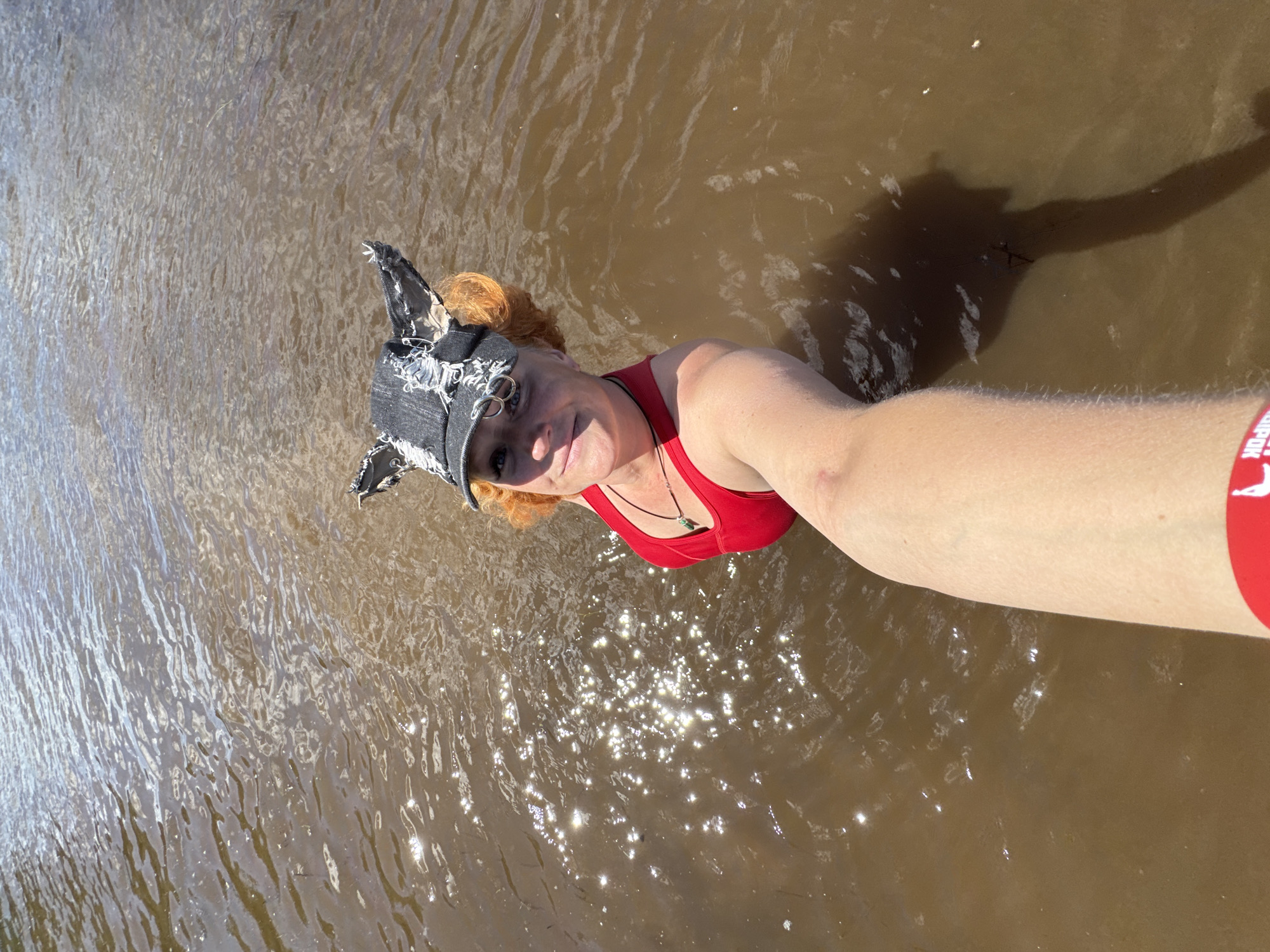 In this self-taken photograph, a woman with vibrant red hair is submerged in murky brown water, likely a river, lake, or pond, given the ripple patterns and sunlight reflecting off the surface. She is wearing a red swimsuit and a dark, distressed baseball cap with decorative silver rings and frayed edges, giving it a unique, edgy appearance. A dark necklace with a green pendant is visible around her neck. Her right arm, extended towards the camera, is bare and slightly tanned, with a red band on her bicep featuring white text. The water appears to be relatively shallow in the foreground, with sunlight creating a shimmering effect. The overall mood seems relaxed and playful, with the woman smiling at the camera. The lighting suggests it is daytime, likely sunny. No specific landmarks are visible to pinpoint the exact location within Chaikovsky, Russia, beyond the presence of a body of water.