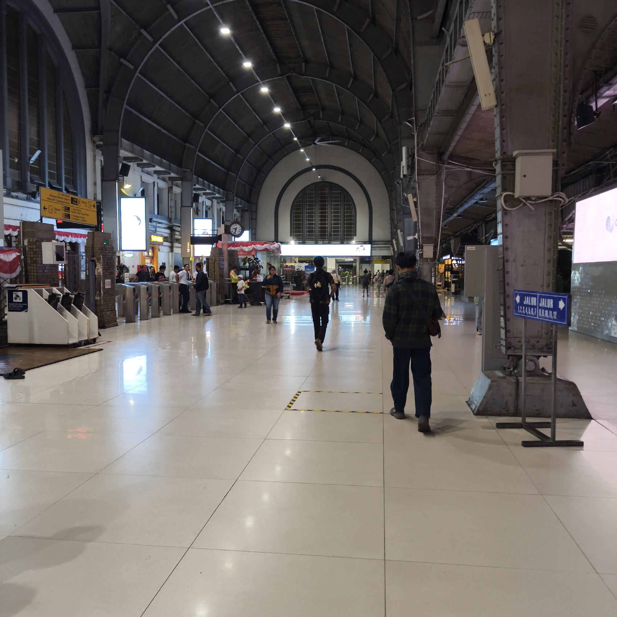 The image depicts the interior of a large, modern train station in West Jakarta, Indonesia. The architecture features a high, arched ceiling with exposed metal beams and numerous lights, suggesting a grand and functional space. The floor is polished and reflective, indicating a clean and well-maintained environment.
In the foreground, a person walks away from the camera, wearing a plaid shirt and dark pants. To the left, a series of turnstiles are visible, with several people in line or passing through them. Behind the turnstiles, a row of yellow signs with Indonesian text provide directional information.
Further back in the concourse, more people are walking and interacting. Several large, illuminated digital displays are mounted on the walls, likely showing train schedules or advertisements. Structural pillars, clad in metal, support the station's roof. One prominent pillar in the mid-ground has a blue sign with white text and arrows indicating "JALUR JALUR 1,2,3,4,5,6,7 8,9,10,11,12", signifying platform numbers.
The overall scene is bustling with activity, typical of a public transportation hub. The lighting is a mix of overhead artificial lights and the illumination from the digital displays. The image captures a moment in the daily operations of the train station.