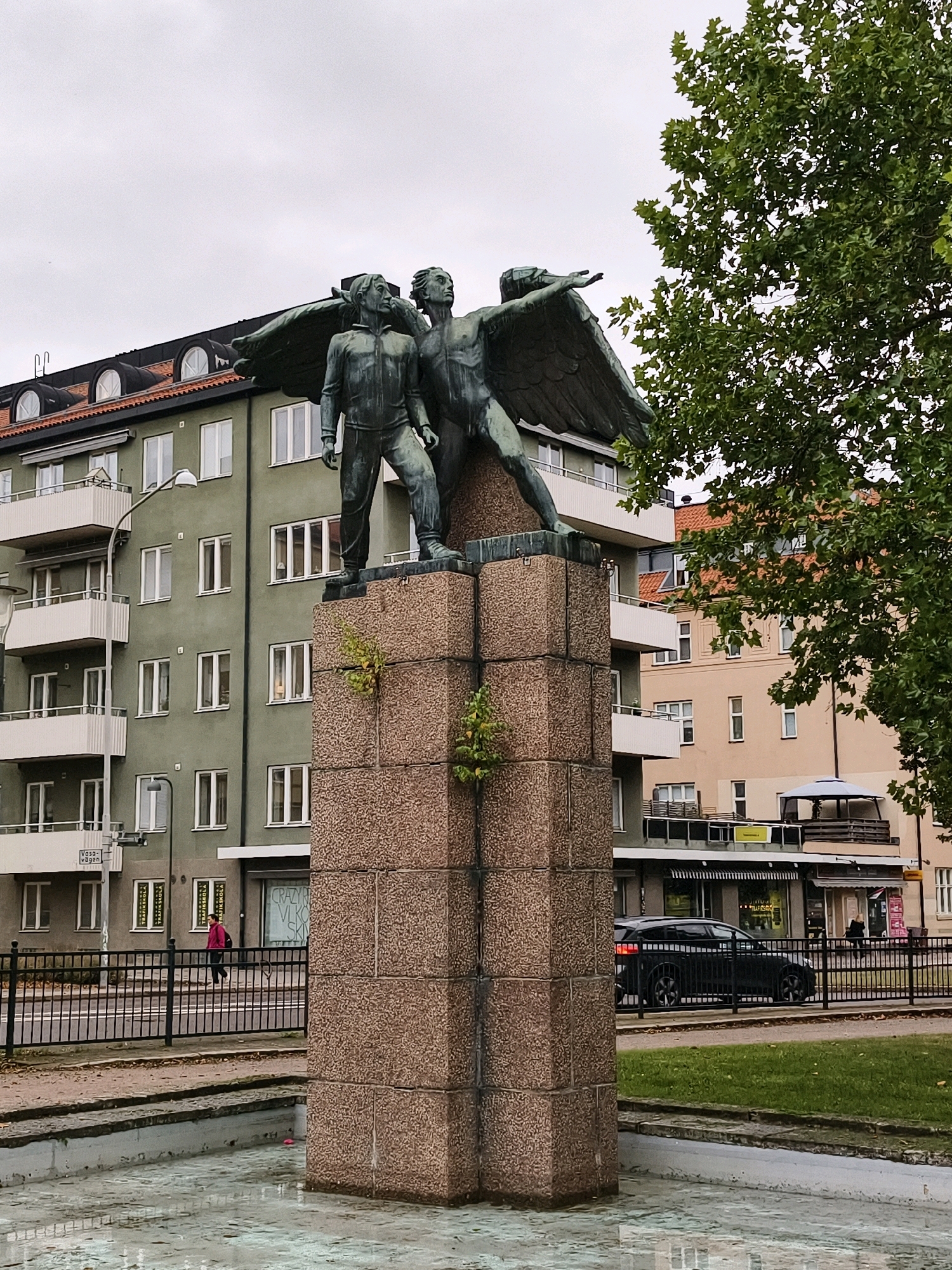A bronze statue of two winged human figures stands on a stone pedestal in an outdoor public space. The figures are positioned side-by-side, with one figure extending an arm outwards and the other looking upwards. Behind the statue, a multi-story green building with white-framed windows and balconies is visible, along with other buildings and a tree with green foliage. A street runs in front of the buildings, with a black metal fence separating the sidewalk from a grassy area. A dark-colored car is parked behind the fence, and a person in a pink jacket walks on the sidewalk. The sky is overcast. This image was taken in Linköping, Sweden, specifically near the public fountain in the courtyard of the former military hospital, which is now part of the Linköping University.