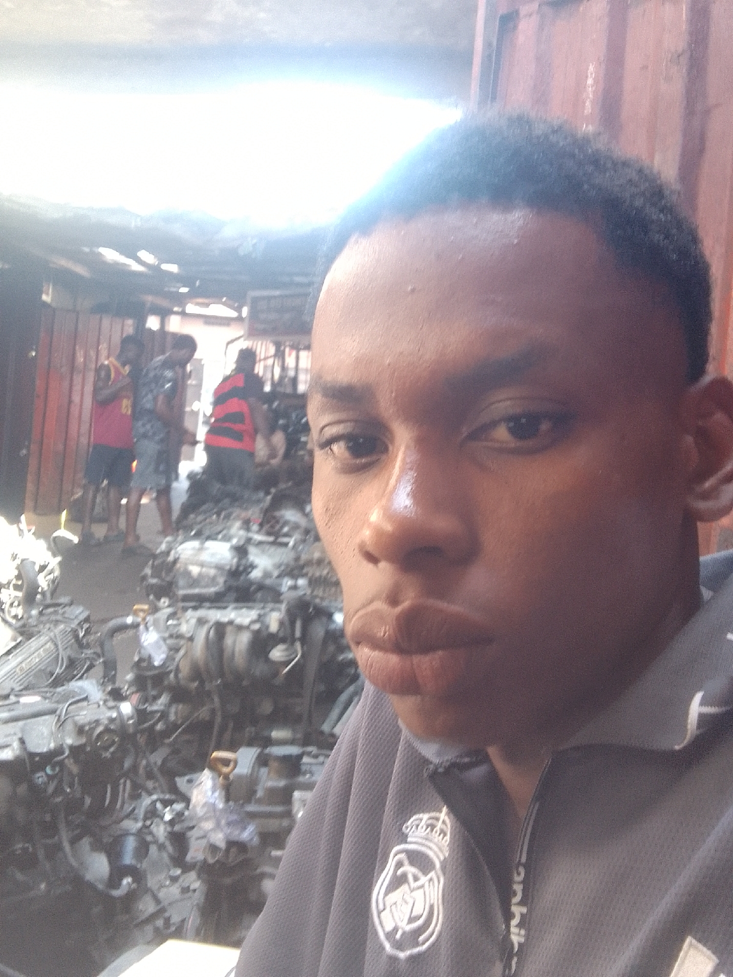 The image shows a close-up selfie of a young man with short dark hair, dark skin, and full lips. He is wearing a grey polo shirt with the Real Madrid logo on the left side and Adidas branding on the collar. In the background, out of focus, is a cluttered scene with several people standing near a large pile of what appears to be used car engines. The setting is likely a marketplace or junkyard in Onitsha, Nigeria, based on the dense arrangement of goods and informal outdoor location. The image is bright, and slightly overexposed.
