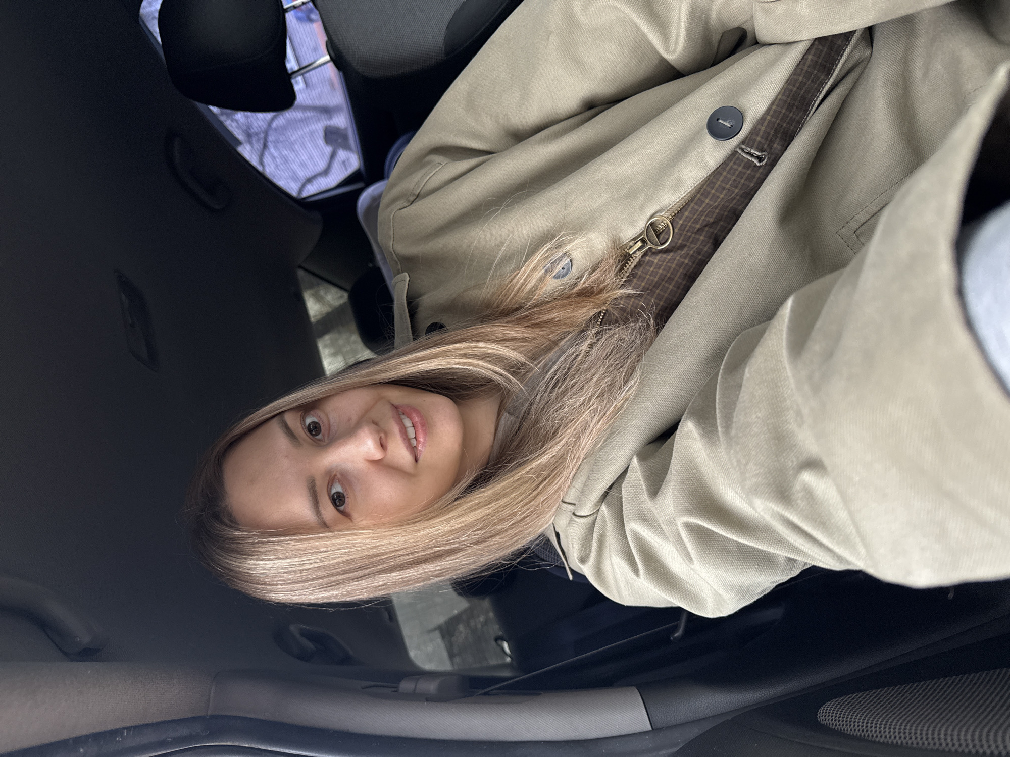 A woman with blonde hair is sitting in a car. She is wearing a light beige jacket with a brown patterned lining visible near the zipper. The car's interior is dark. Outside, there is a glimpse of a street scene with buildings and fences. The exact location in Chaikovsky, Russia, cannot be determined from this image alone.