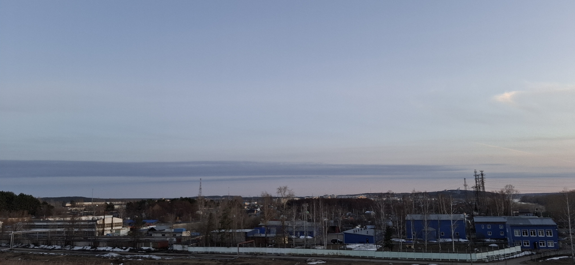 A daytime view of a city with a blue sky and a layer of clouds. In the foreground is a fence, followed by a rail. Behind the fence are several buildings painted blue. In the distance, there is a wooded area and a tower. The photo was taken in Chaikovsky, Russia.