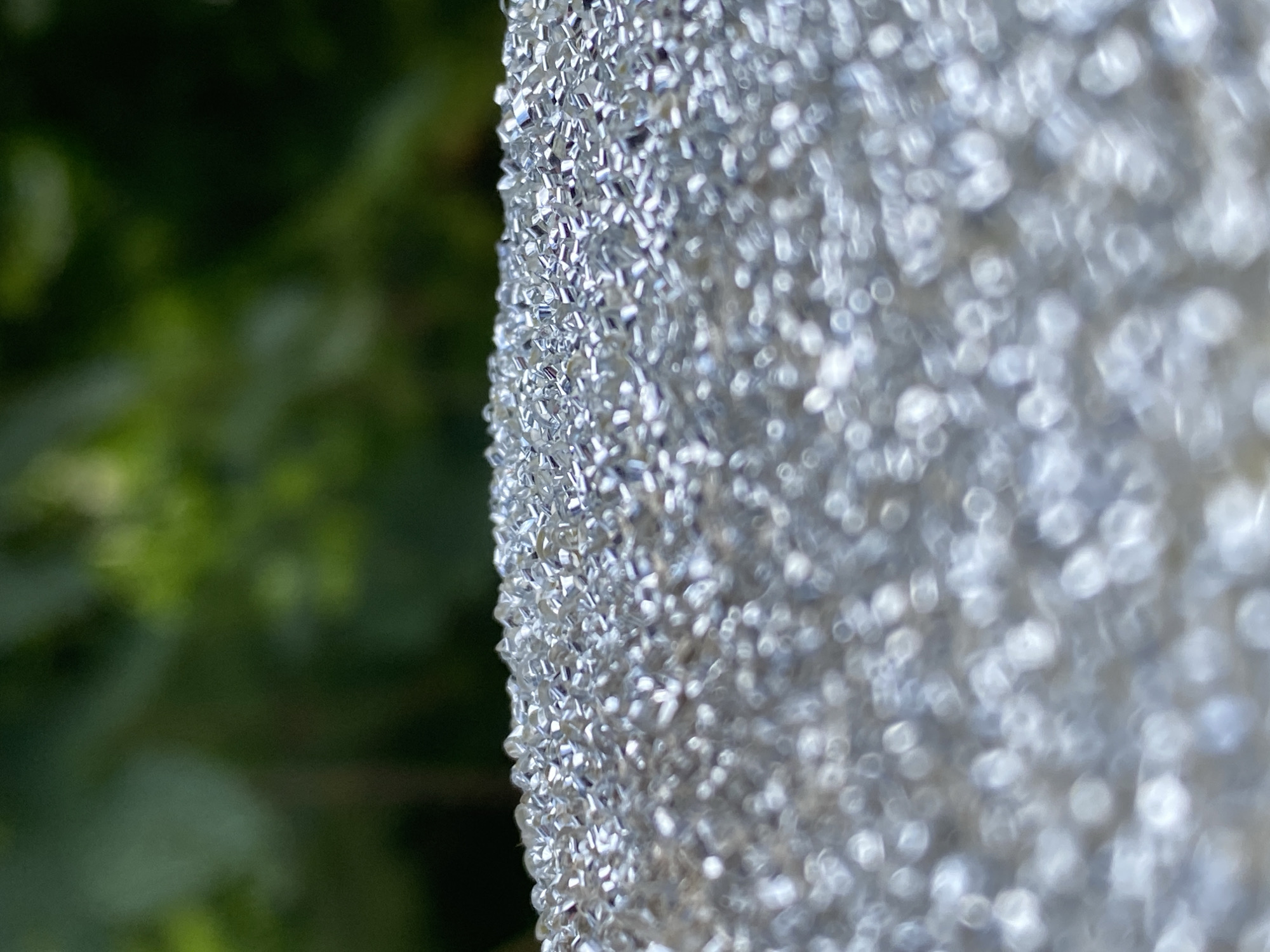 The image is a close-up, shallow depth-of-field shot focusing on a surface covered in small, reflective, crystalline or bead-like elements. The background is blurred and appears to be foliage in shades of green and dark green, suggesting an outdoor environment, possibly a garden or park. The crystalline surface dominates the foreground, with individual elements visible and reflecting light, creating a shimmering effect. The background bokeh blurs any discernible details of the foliage. There are no people, actions, or interactions visible in the image. The scene depicts an abstract texture against a natural backdrop. The location in Purkersdorf, Austria, is not evident from the visual information provided.