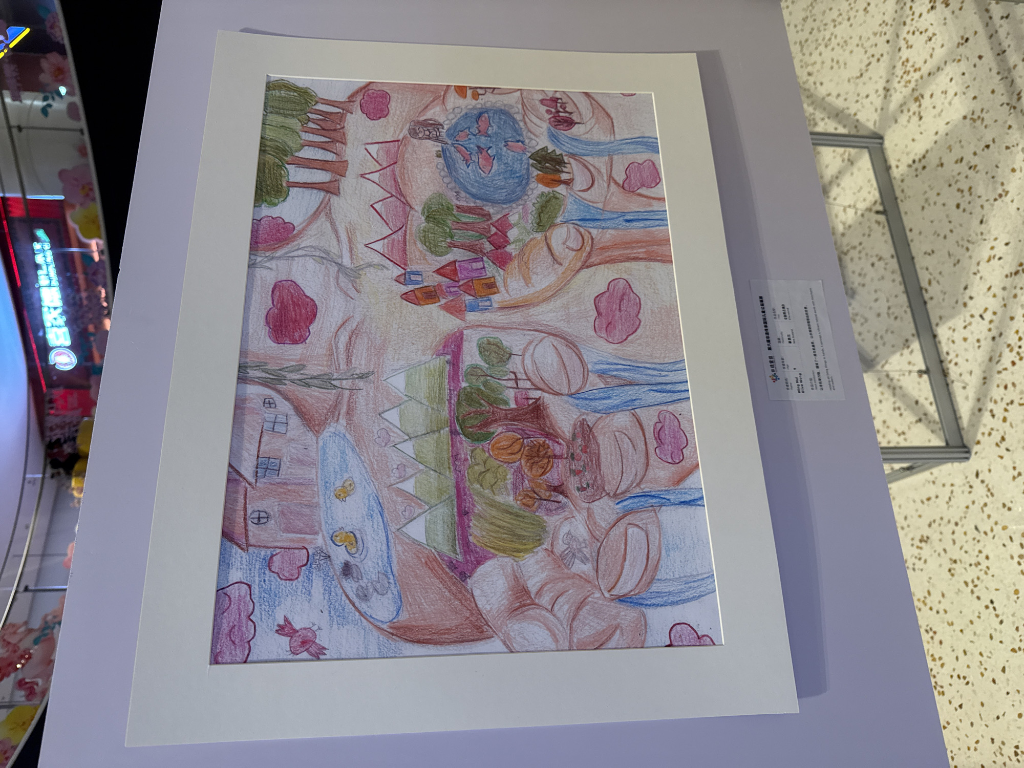 This image, captured in Haidian District, China, displays a framed children's drawing presented on a light purple exhibition panel. The drawing, rendered in colored pencils, depicts a fantastical and vibrant landscape. The composition is notable for its use of rounded, skin-toned shapes, suggesting hands or organic forms that cradle various scenes.
In the upper left, two brown houses with windows and cross-hatched doors stand beside a pond where two yellow ducks swim. A red bird with a distinctive wing pattern flies nearby under pink, cloud-like formations. The upper right features a cluster of green trees with brown trunks. In the mid-ground, jagged green mountains with white peaks are visible, bordering a section where colorful houses with red roofs are clustered. Below this, a pond contains several red fish, and a small, brown-haired figure is depicted near its edge. Blue waterfalls cascade downwards from multiple points, integrating with the organic landscape shapes and flowing into lower sections of the drawing. These lower sections also feature more trees, foliage, and additional pink, cloud-like forms.
The drawing is encased in a simple white frame with a white mat. Below the artwork, a small rectangular white plaque displays black text, providing details about the artwork and its creator, including the artist's name "Wing Mui Olive" and the artwork title "自然之境" (Nature's Realm). The exhibition panel is supported by a minimalist metal stand visible at the bottom right.
The background, largely out of focus, reveals elements of an indoor commercial environment, likely a shopping mall. Neon signs with Chinese characters, including "白天鵝 BLACK," are visible, suggesting retail or dining establishments. Decorative floral elements are present on a railing or upper display structure. The floor features a light-colored terrazzo or speckled tile pattern.