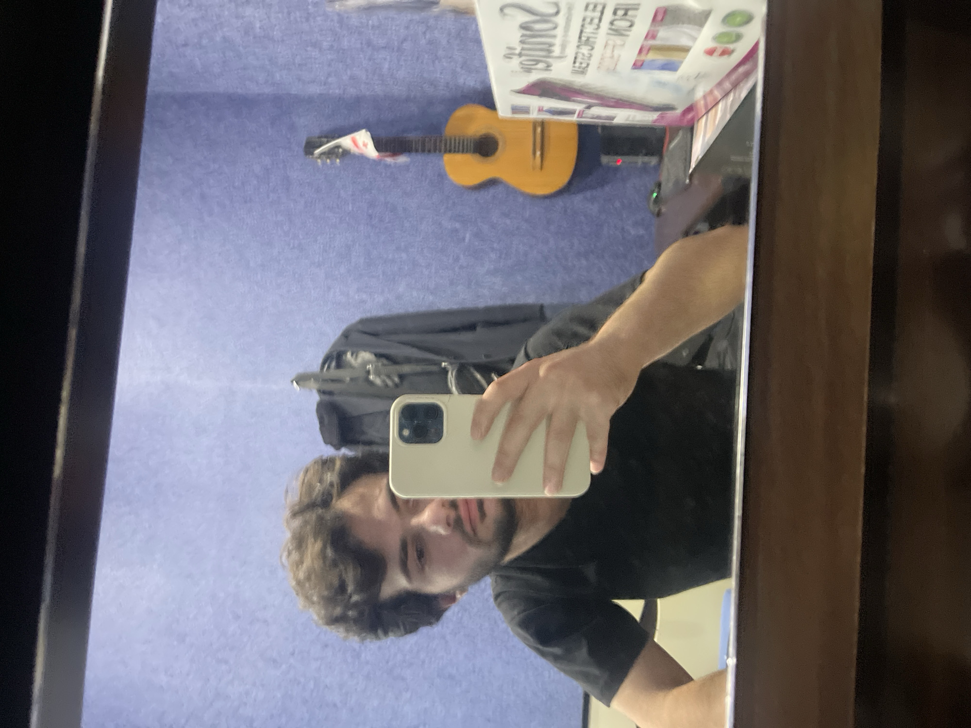 A man takes a mirror selfie in what appears to be a recording studio. He is wearing a black t-shirt and holding a white iPhone in his right hand. The background shows a purple textured wall, a guitar with a white cloth hanging from its headstock, and a coat hanging on a rack. A product box with colorful images and text is visible on a table in the foreground. The scene is illuminated by artificial light.
