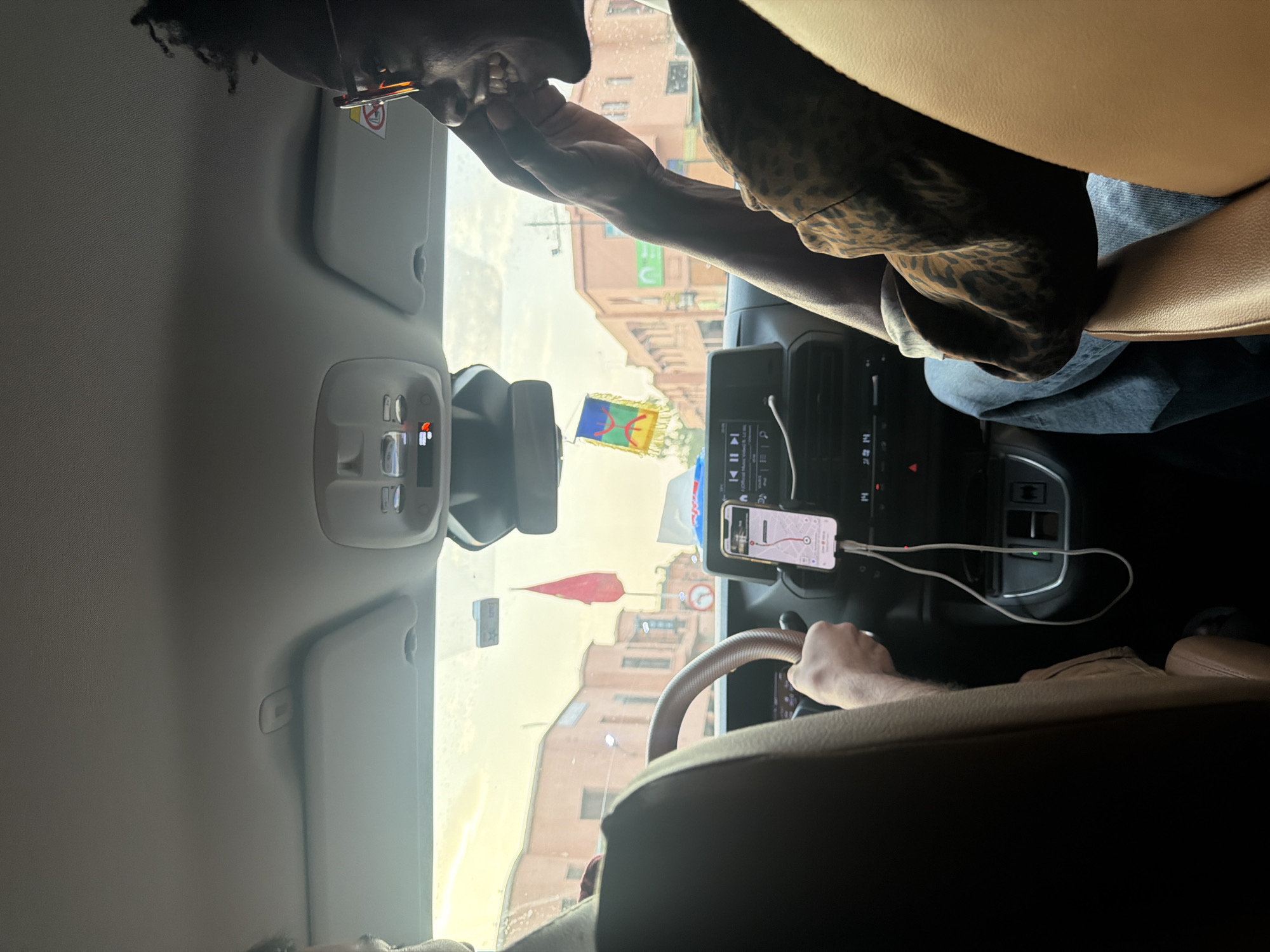 The image shows the interior of a car in Marrakesh, Morocco, with two people inside. The driver, whose arm is visible gripping the steering wheel, is focused on the road. In the passenger seat, a Black man wearing glasses and a patterned shirt is smiling and looking towards the right. Outside the car, traditional Moroccan buildings with orange facades and arched windows can be seen under a hazy sky. A red flag is visible on a pole outside the car, and a colorful flag hangs from the car's rearview mirror. A smartphone mounted on the dashboard displays a navigation app. The scene suggests a typical street view within the city of Marrakesh.