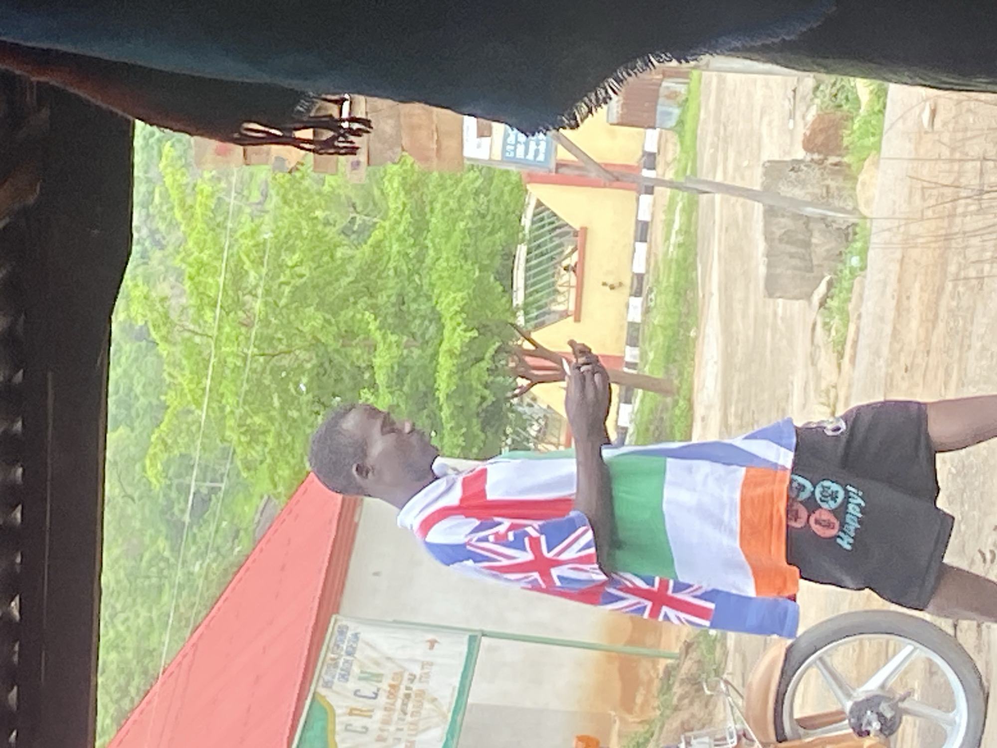 A man stands in an outdoor setting in Mararaba, Nigeria. He wears a shirt designed to resemble the flags of the United Kingdom, Ireland, and India, along with dark-colored shorts imprinted with the word "Happy!" and circles. He is holding a small object in his hands, possibly using a mobile device. In the background, there are green trees, buildings with red roofs and light-colored walls, a white sign board, and a bicycle wheel.