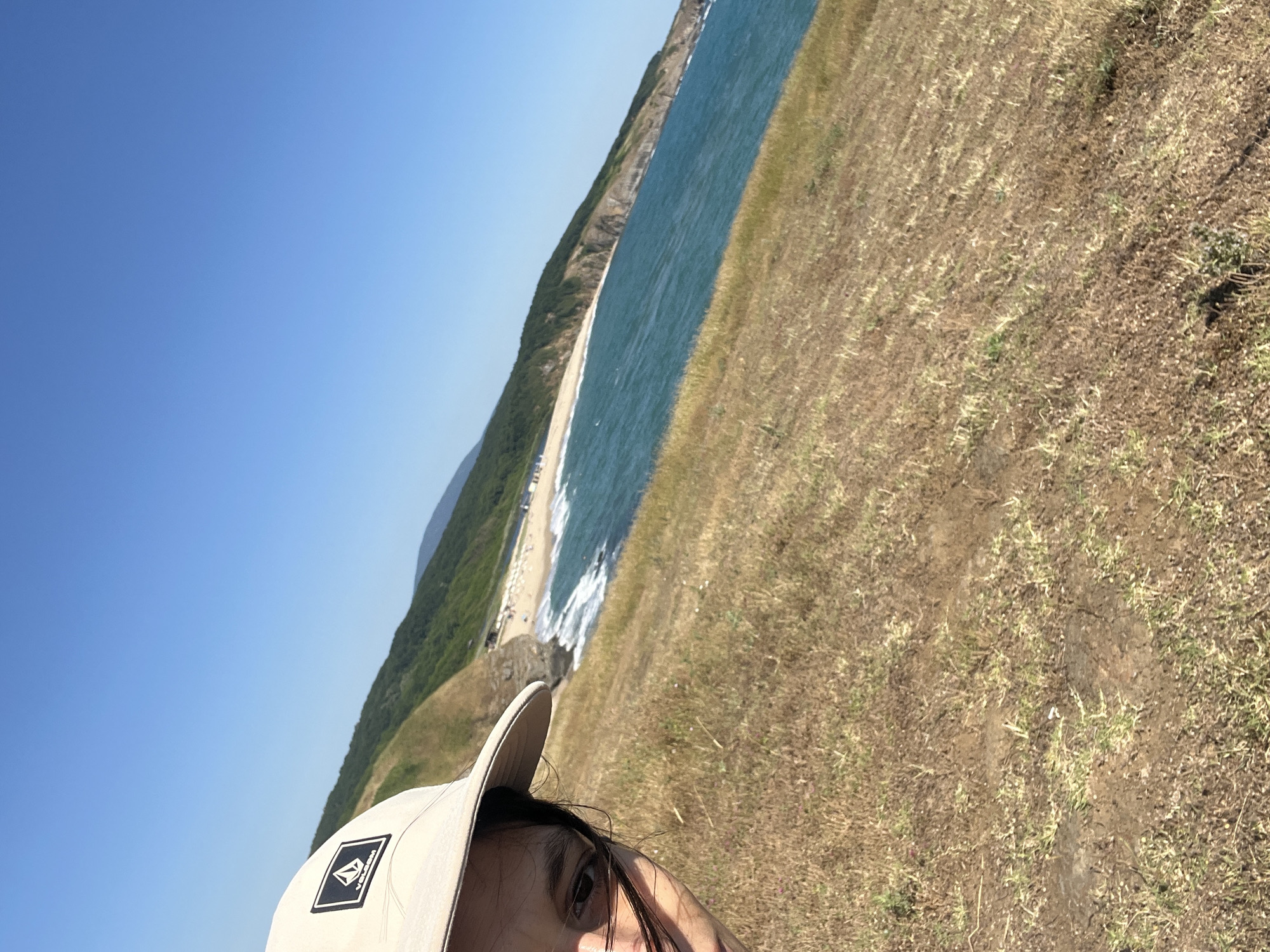 An eye-level, selfie-style shot shows a person wearing a beige baseball cap with a black and white Volcom logo, with their face partially visible in the left foreground, looking to the right. They are on a grassy, sun-drenched hillside covered in dry, yellowed grass and scattered small green shoots. The background shows a bright blue sky with no clouds. Below the hillside, a sandy beach with a few scattered umbrellas and people is visible, bordered by turquoise water with white waves. The coastline curves around, with a steep, green, tree-covered hill rising behind the beach and continuing into the distance. The image appears to be taken in the Sinemorets area of Bulgaria, likely near Veleka Beach or Silistar Beach, known for their scenic coastal landscapes.