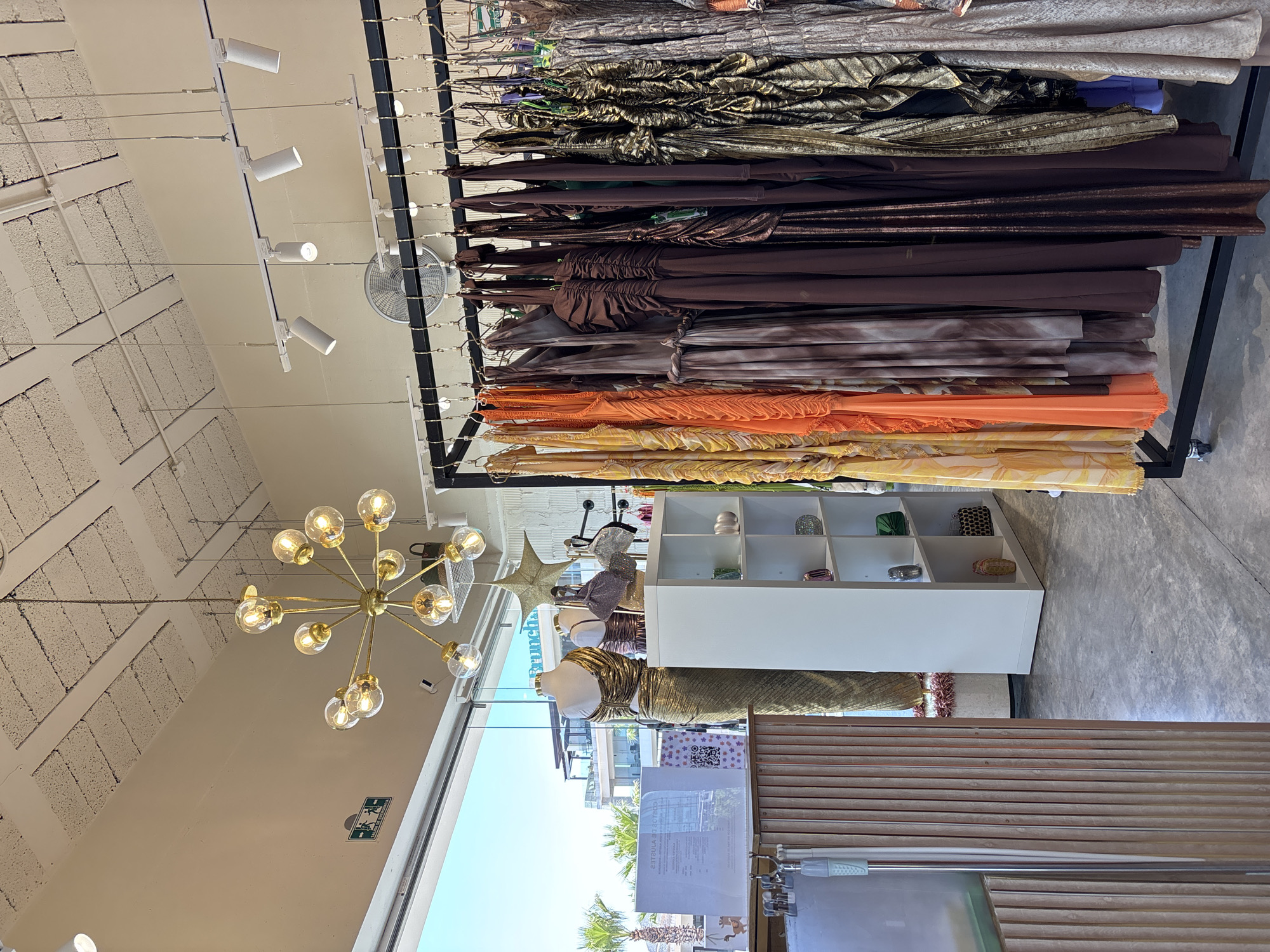 This media file presents an interior view of a contemporary clothing store or boutique, likely situated in San Bernardino Tlaxcalancingo, Mexico, as evidenced by the clear sky and palm trees visible outside. The image captures a bright, daytime scene, illuminated by ample natural light streaming through a large window on the right.
The primary subjects are numerous garments, predominantly dresses or formal wear, hanging neatly on a central display rack. The collection features a palette of warm, earthy colors, including various shades of gold, bronze, dark brown, orange, and yellow, with some fabrics exhibiting metallic finishes or ruched textures. Towards the lower right, at least two mannequins are visible, with one prominently dressed in a shimmering gold, possibly pleated, outfit.
The store's interior design is modern, characterized by a grey concrete floor and light-colored walls and ceiling. The ceiling incorporates both areas of exposed concrete blocks and smooth, painted sections. Illumination is provided by several white track lighting fixtures mounted on the ceiling, as well as a striking brass-colored Sputnik-style chandelier with multiple clear, spherical bulbs. A circular ceiling fan is also visible in the upper right quadrant.
Next to the large window, a white shelving unit displays small accessories like purses or clutches. Beyond the window, the outdoor scene reveals a clear blue sky, vibrant green palm trees, and glimpses of other buildings, indicative of a warm, sunny climate.
Visible text includes a partially obscured white sign near the window, which appears to be a "NOTICE" (likely "AVISO" in Spanish) accompanied by a QR code. A small green exit sign with white pictograms is also mounted on the wall below the chandelier. The overall impression is one of a clean, well-lit, and spacious retail environment.