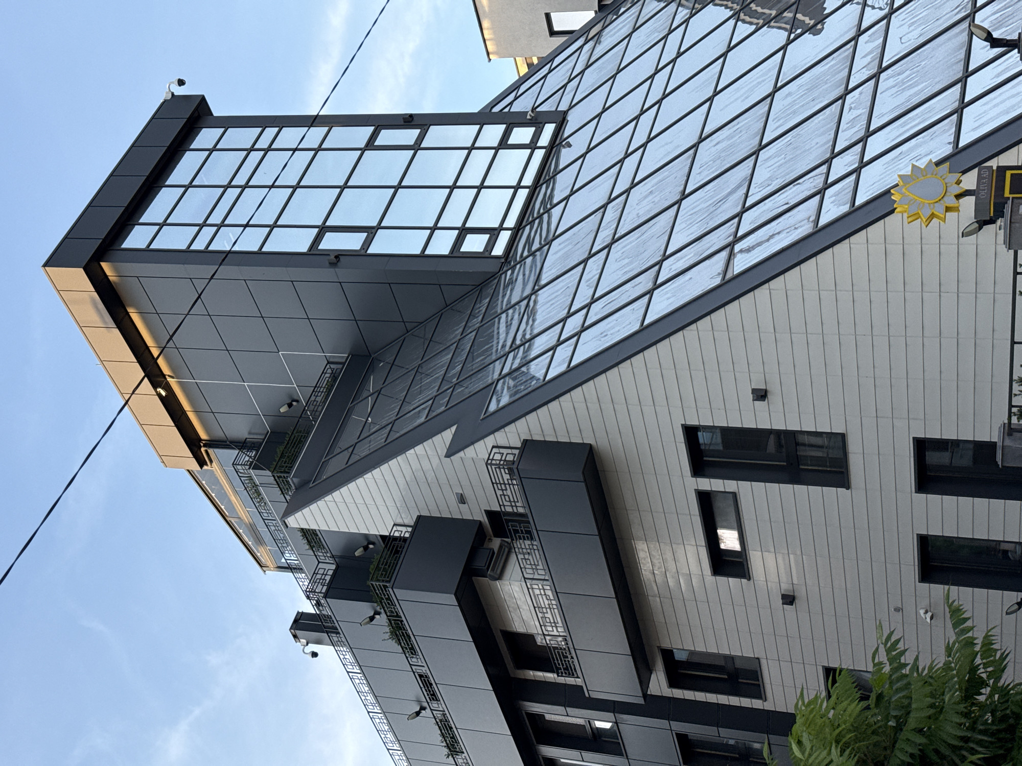 The image shows the exterior of a modern, multi-story building. The building features a geometric design with a prominent glass facade composed of square panes. The building's facade is also composed of gray and white panels. Some balconies are visible on the lower floors. Security cameras are mounted on the exterior. A yellow decorative sun-shaped sign is attached to the side. The sky is visible in the background. The location is likely Sofia, Bulgaria. Vegetation is partially visible at the bottom of the image. Electrical wiring is partially visible.