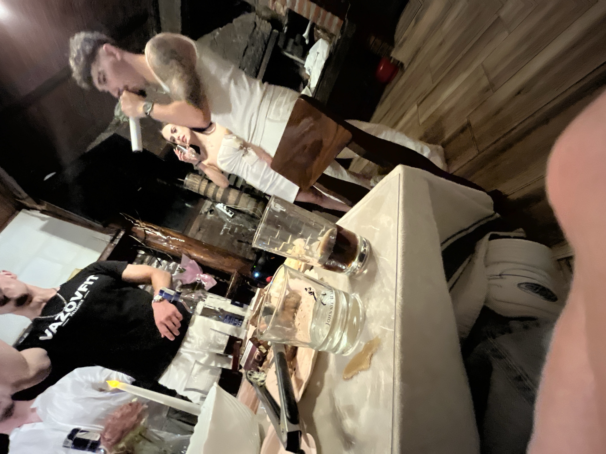 The image shows an indoor scene with multiple individuals. A man with short, dark hair and tattoos is holding a microphone and speaking. A woman is standing near him, holding a glass. Another man in a black t-shirt with "VAZOVFIT XLLENCE" written on it is also present. A table is in the foreground with glasses, one of which is a "JOHNNIE WALKER" branded glass. There are also plates and cutlery on the table. The background suggests a dimly lit indoor space with wood paneling and what appears to be a fireplace. The exact location in Presoka, Bulgaria, is unclear based on the image. The actions suggest a social gathering or performance.