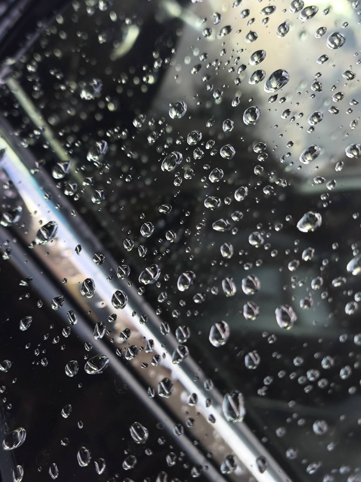 This is a close-up, abstract photograph of water droplets on a dark, reflective surface, likely a window or glass panel. The droplets vary in size and shape, with some appearing perfectly round and others elongated or irregular. They are illuminated from above and to the left, creating bright highlights and casting subtle shadows. The background is a blur of dark grays, blues, and faint iridescent tones, suggesting a dimly lit environment or the reflection of ambient light. The overall impression is one of a rainy day, with the water distorting and magnifying the reflections of the surroundings. The image does not contain any people, specific identifiable objects, or actions. There is no information within the image itself to suggest a location in Kaliningrad, Russia.