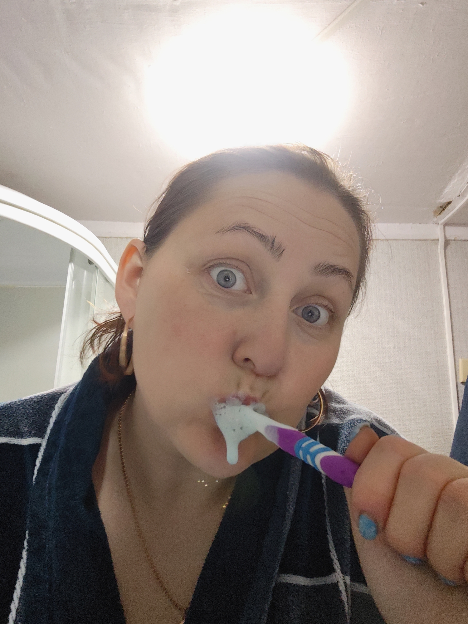 A woman is in a bathroom, brushing her teeth. She is wearing a blue and white plaid robe and gold hoop earrings and a thin gold chain. The toothpaste is blue. The toothbrush has a purple and white handle. She is looking at the camera with wide eyes. The bathroom has a white ceiling and walls. A shower stall is visible on the left. The location is Фоки, Russia.
