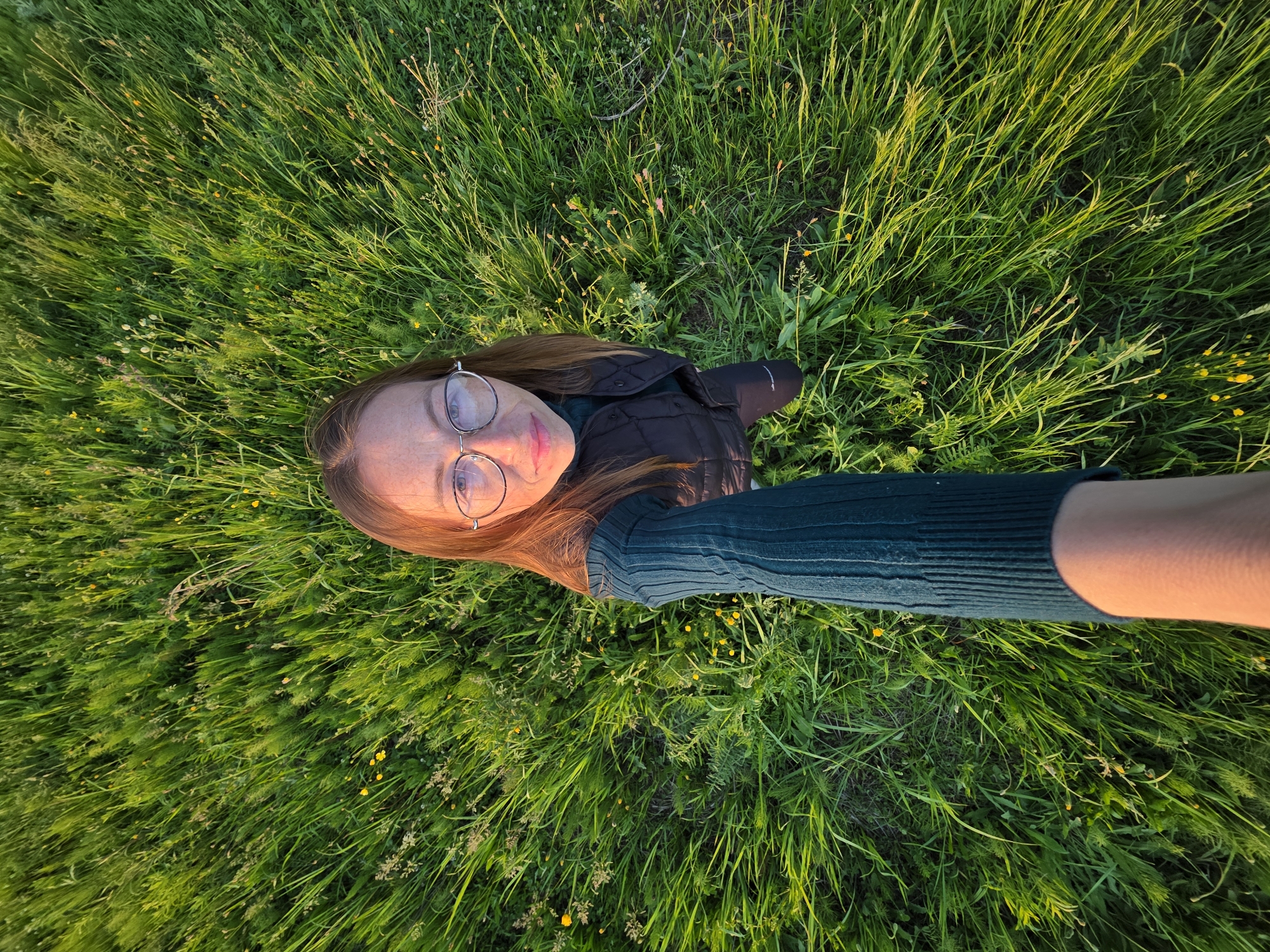 An overhead, wide-angle shot captures a woman lying in a field of lush green grass, illuminated by the warm, golden light of what appears to be late afternoon or early evening. The camera is positioned directly above her, likely held by her extended arm, giving the impression of a selfie taken from a bird's-eye view.
The woman has reddish-brown hair and wears round, silver-rimmed glasses. Her face, visible at the top left of the frame, shows a calm expression with a slight, gentle smile. She is dressed in a dark, possibly black or brown, puffer jacket over a dark green, ribbed long-sleeved top. The golden sunlight casts a warm glow on her skin and the top of her arm, highlighting the vibrant colors.
The setting is a natural landscape, likely a meadow or park in Chaikovsky, Russia, characterized by abundant, tall green grass interspersed with small yellow wildflowers. The overall atmosphere is peaceful and serene, emphasizing a moment of relaxation in nature on a sunny day. No text is visible in the image.
