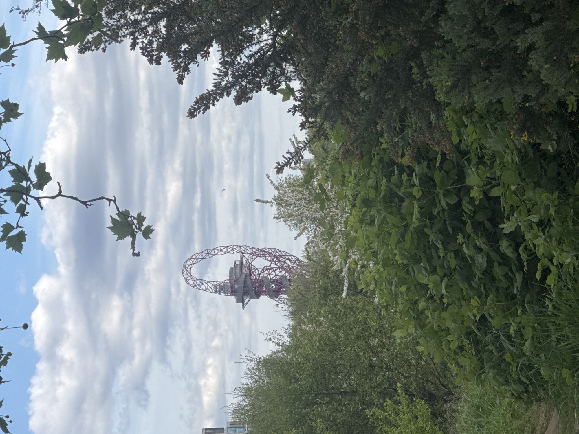 The image captures a view of the ArcelorMittal Orbit, a sculpture and observation tower located in the Queen Elizabeth Olympic Park in Stratford, London, United Kingdom. The Orbit is partially obscured by trees and foliage in the foreground. The sky is overcast with clouds. There are no people visible in the image. A few buildings are seen at the bottom left corner. A bird is flying at the center left.