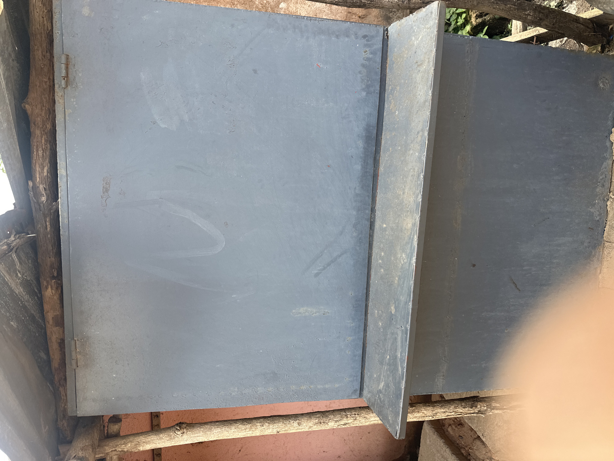 An outdoor, medium shot shows a gray, rectangular storage container with a hinged lid. The lid is slightly ajar and rests on a wooden support structure. The container is situated in an outdoor environment with a pink wall visible to the left and foliage to the right. The overall scene appears to be in a state of disrepair, with rust present on the hinges of the lid. The storage container is elevated on a lower, similar-shaped base.