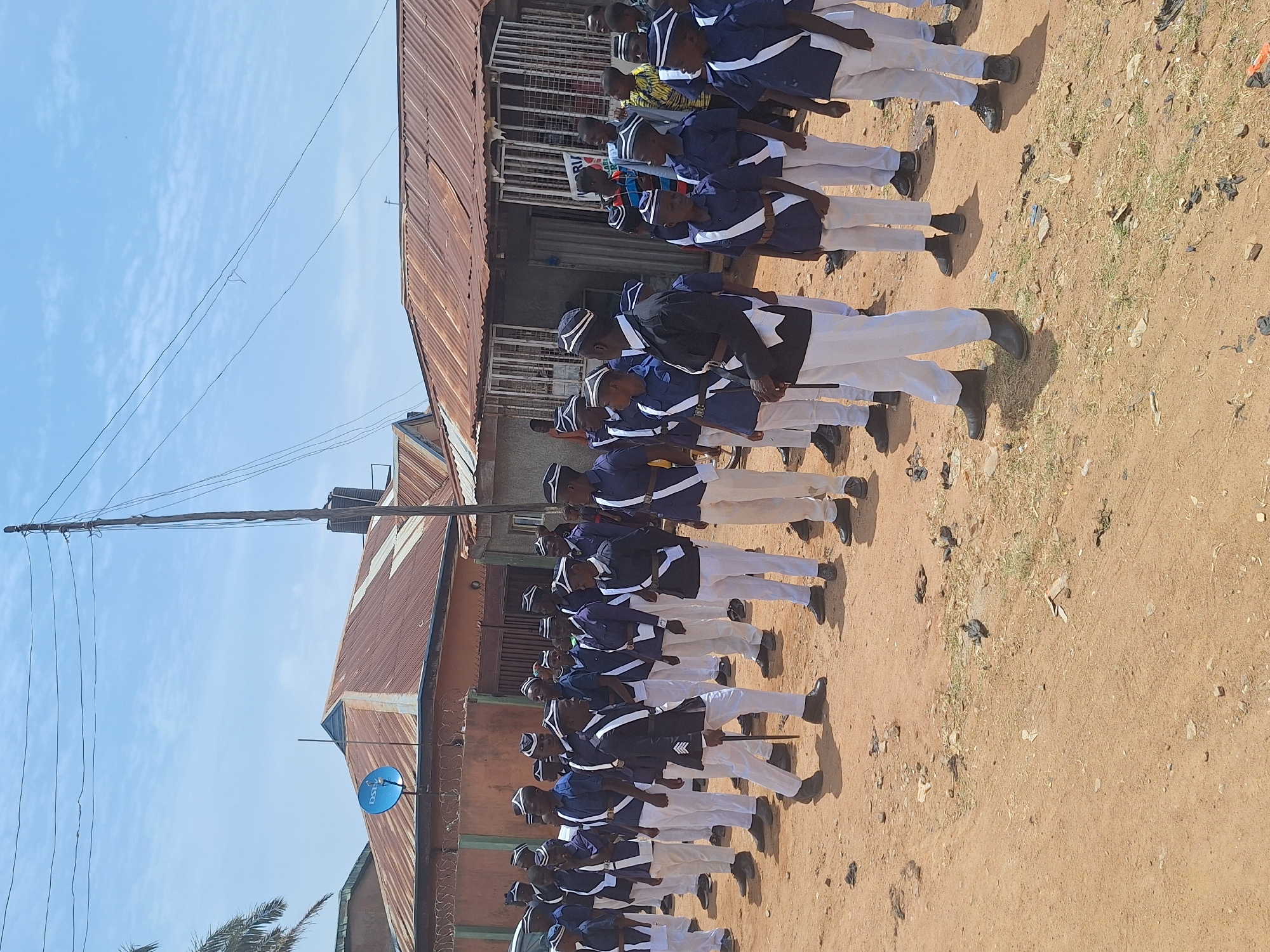 A group of people in matching uniforms, with dark blue jackets and white trousers, are lined up on a dirt road. They are arranged in a column, with each person standing close to the next. A corrugated metal building with a satellite dish is on the left of the frame. The location is likely Sabon Tasha, Nigeria, based on the context provided.
