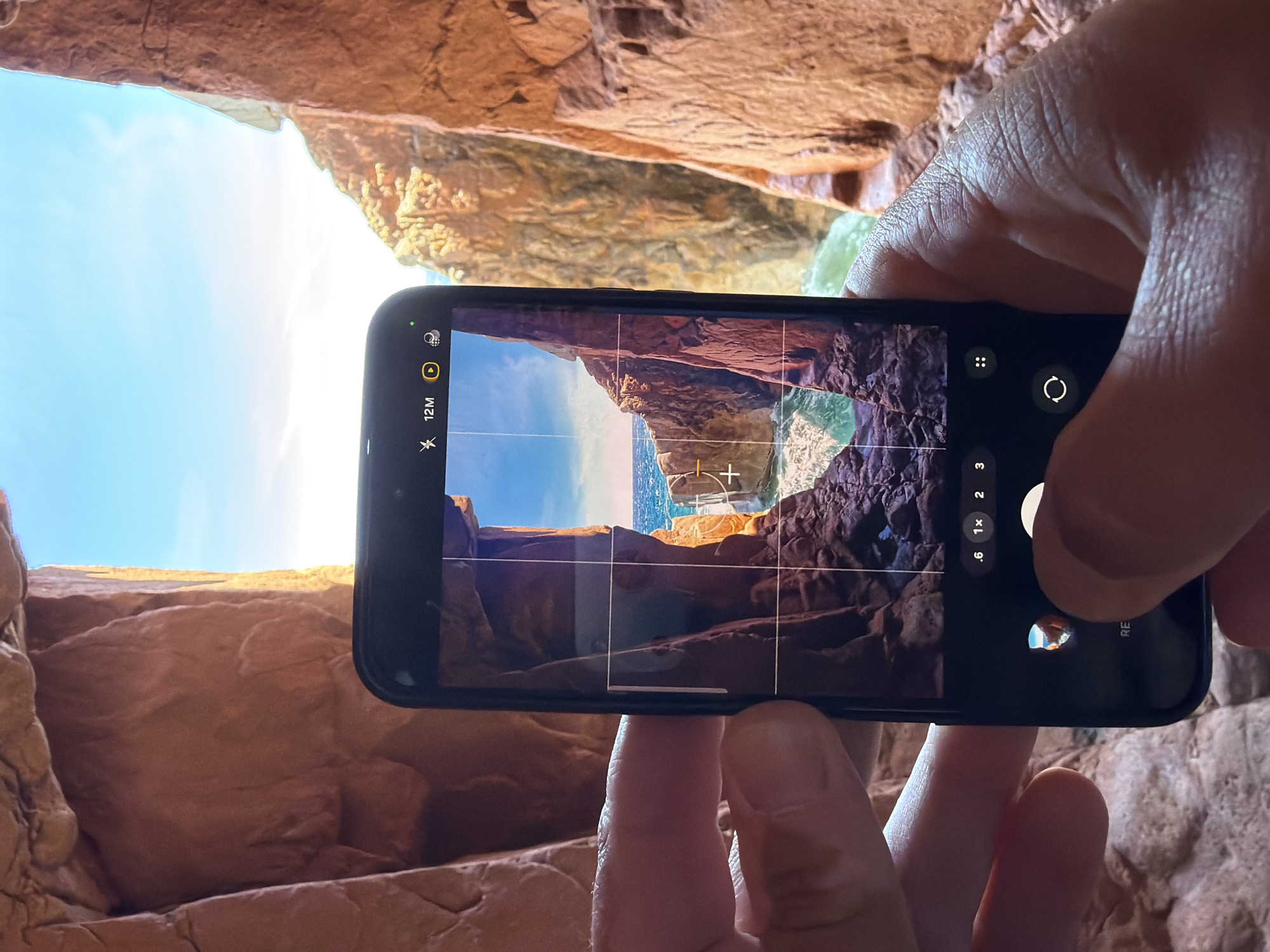 An outdoor, eye-level, medium shot captures a person's hands holding a smartphone to take a photo. The phone is positioned in the center of the frame, angled slightly towards the viewer, and displays a scene of rocky cliffs and the ocean. The person's hands are on either side of the phone, with their thumbs and fingers clearly visible. The phone screen shows a grid overlay, suggesting the camera app is active and the user is framing a shot. In the background, the rough, textured surfaces of reddish-brown rock formations create a cave-like environment. Sunlight streams in from an opening, illuminating the rocks and the distant ocean, which is a vibrant blue with white waves. The sky above is a clear, bright blue. The location is the sea caves at Cala Mariolu on the coast of Baunei, Sardinia, which is part of the municipality of Tortolì, Italy.