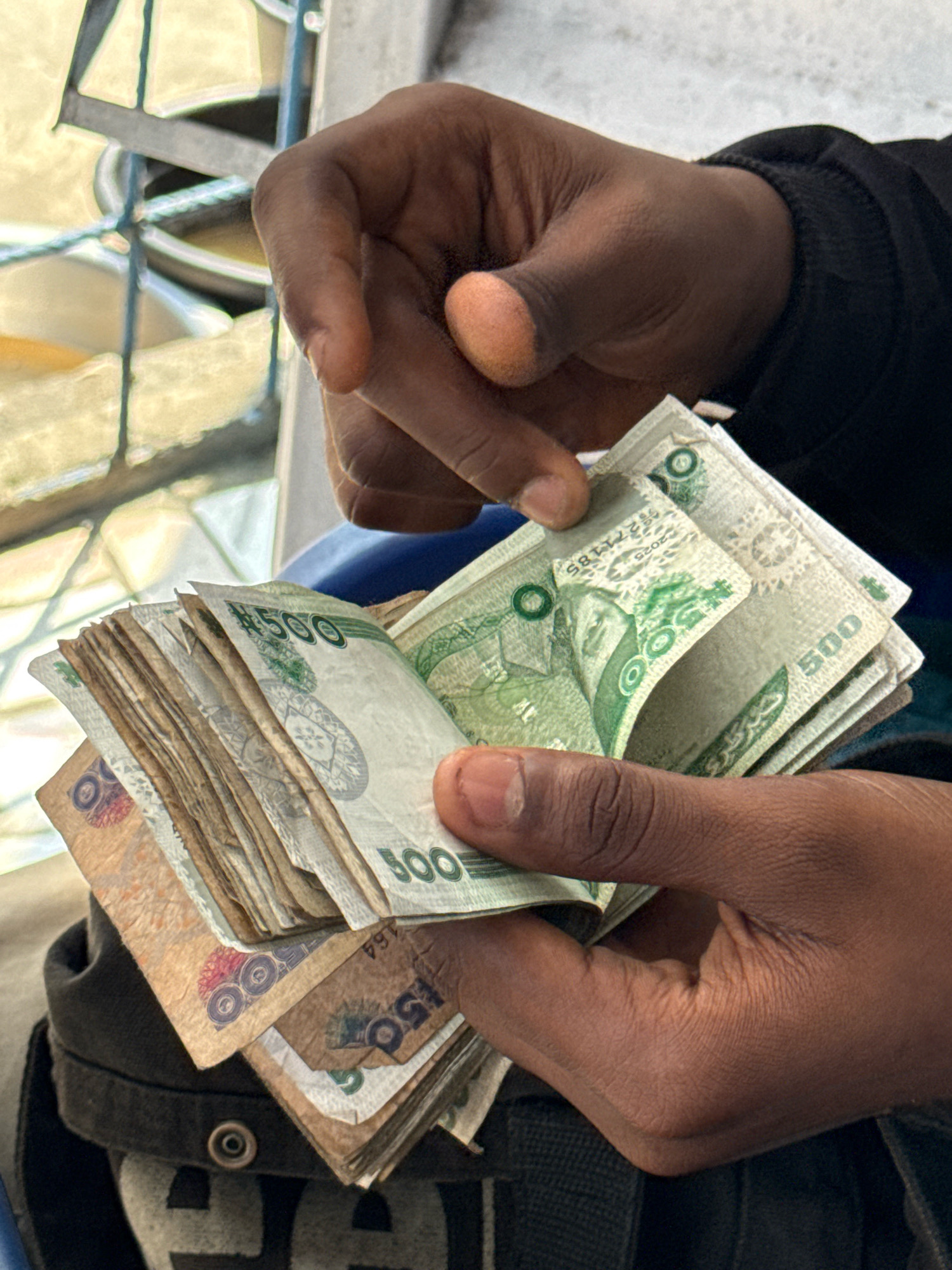 A close-up shot shows a person holding a wad of cash. The money appears to be Nigerian Naira, with denominations of 500 and 150 visible on some bills. The bills are predominantly green and brown. The person's hands are dark-skinned, and they are wearing a black jacket. The background is out of focus, but appears to be an outdoor setting with some metal bars and light-colored surfaces, possibly suggesting a market or street scene. There is no indication of the specific location within Ado, Nigeria.