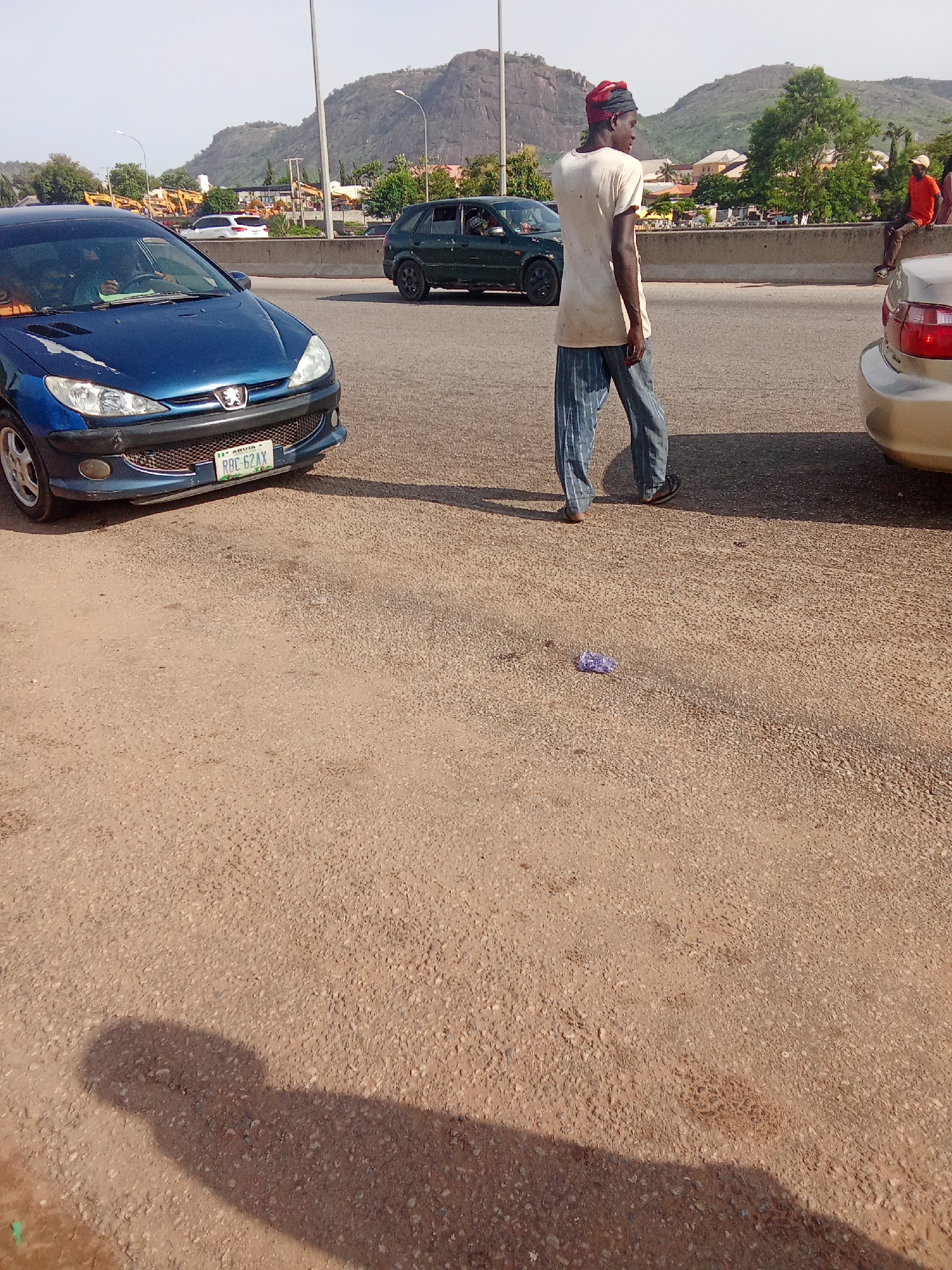 A man wearing a white t-shirt, striped pants, and a red headscarf stands on the side of a road. He is looking towards the right of the image. A blue car is parked to his left, and a portion of a gold car is visible to his right. In the background, there is a concrete barrier, some trees, buildings, and mountains under a clear sky. The ground is covered in gravel and dirt.