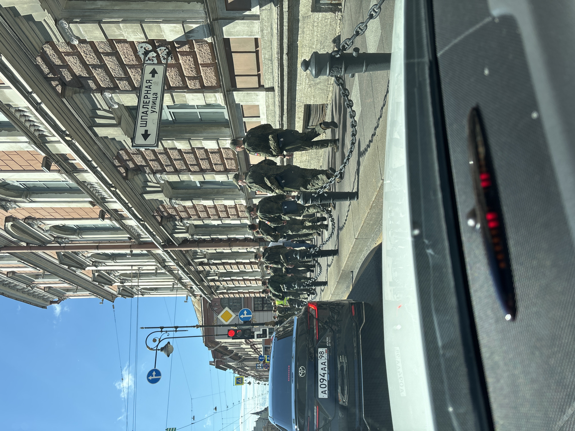 The image shows a street scene in Saint Petersburg, Russia, on a sunny day. The view is from inside a vehicle, looking forward. In the foreground, the dashboard of the vehicle is visible. In the middle ground, a black car with a Russian license plate (A094AA 98) is stopped in traffic. Beyond the car, a group of soldiers in camouflage uniforms are walking along a sidewalk. They are walking in the same direction as the car. The sidewalk is bordered by a decorative chain fence. In the background, there is a large, ornate building with a brick facade and arched windows, typical of Saint Petersburg architecture. A street sign with Russian text and arrows is mounted on the building. Traffic lights and other street signs are also visible. The sky is blue with a few wispy clouds.