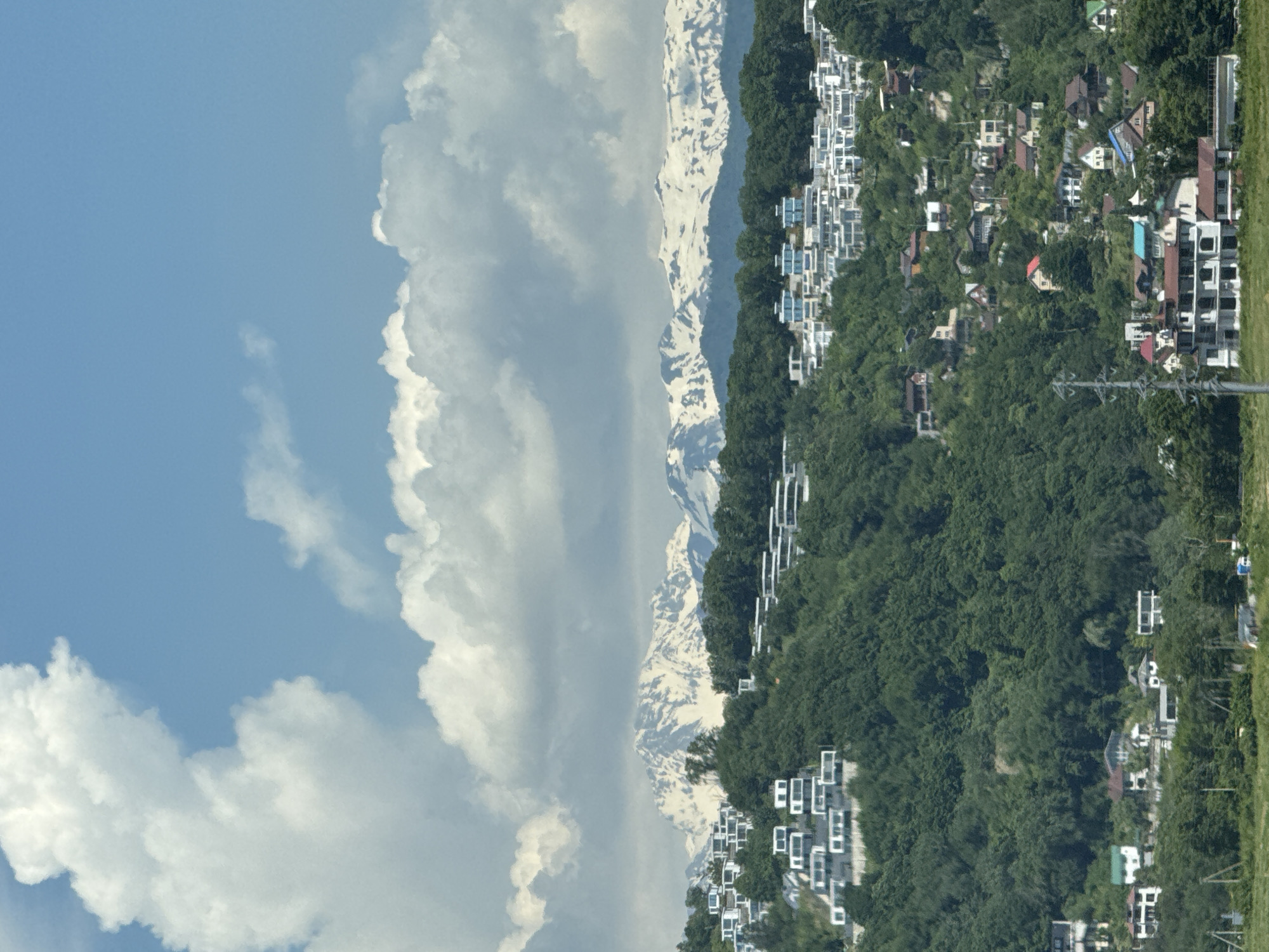 This detailed image depicts a panoramic landscape overlooking a mountainous and hilly region, characteristic of the Greater Sochi area in Russia, with the snow-capped Caucasus Mountains in the background. The upper portion of the frame features a bright blue sky partially obscured by large, fluffy white cumulus clouds, indicating a clear and sunny day.
The horizon line is dominated by a majestic range of distant mountains, identifiable as the Caucasus Mountains, with significant patches of snow on their peaks and upper slopes. Below the mountain range, lush green hills covered in dense deciduous and coniferous forests descend into the mid-ground and foreground.
Scattered across these verdant hillsides are numerous residential buildings. In the upper-mid ground, particularly on the left and right slopes, are clusters of modern, minimalist houses characterized by white facades and flat roofs, often arranged in terraced configurations. Further down the slopes and into the foreground, a mix of older and newer residential structures is visible, with varying architectural styles and roof colors, including red, brown, and blue. Some larger, multi-story buildings are present closer to the bottom of the frame. A prominent utility pole with visible power lines stands vertically in the lower-right foreground. No individuals or explicit actions are discernible within the frame. The overall scene suggests a developed residential area integrated into a natural, mountainous environment.