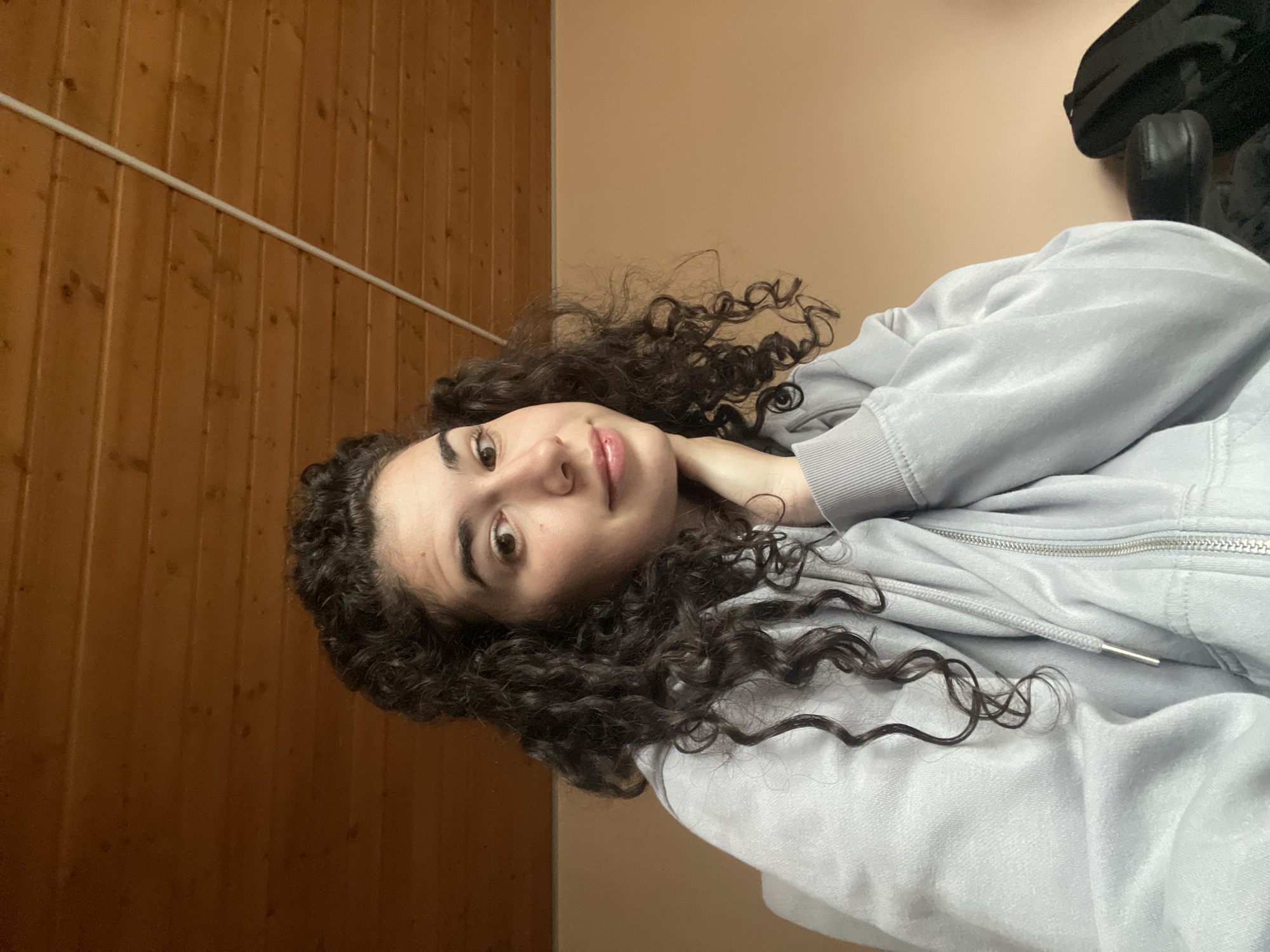 The image displays a head-and-shoulders portrait of a young woman, likely taken indoors. She has dark, voluminous curly hair and is looking directly at the camera with a neutral to slight smile. She is wearing a light grey hooded sweatshirt with a visible zipper and drawstrings. Her left hand is subtly placed near her neck or shoulder.
The background is divided into two main sections: on the left side, there is a wooden paneled wall with vertical planks, across which a white horizontal pipe or conduit runs. On the right side, behind her head, is a plain, light beige or tan-colored wall. A dark object, possibly a bag or part of a chair, is partially visible in the upper right background. The lighting is soft and diffuse, indicating an indoor environment without strong shadows. The overall scene suggests a casual and relaxed setting, possibly a residential room. There is no visible text in the image. The location is contextualized as Sofia, Bulgaria.