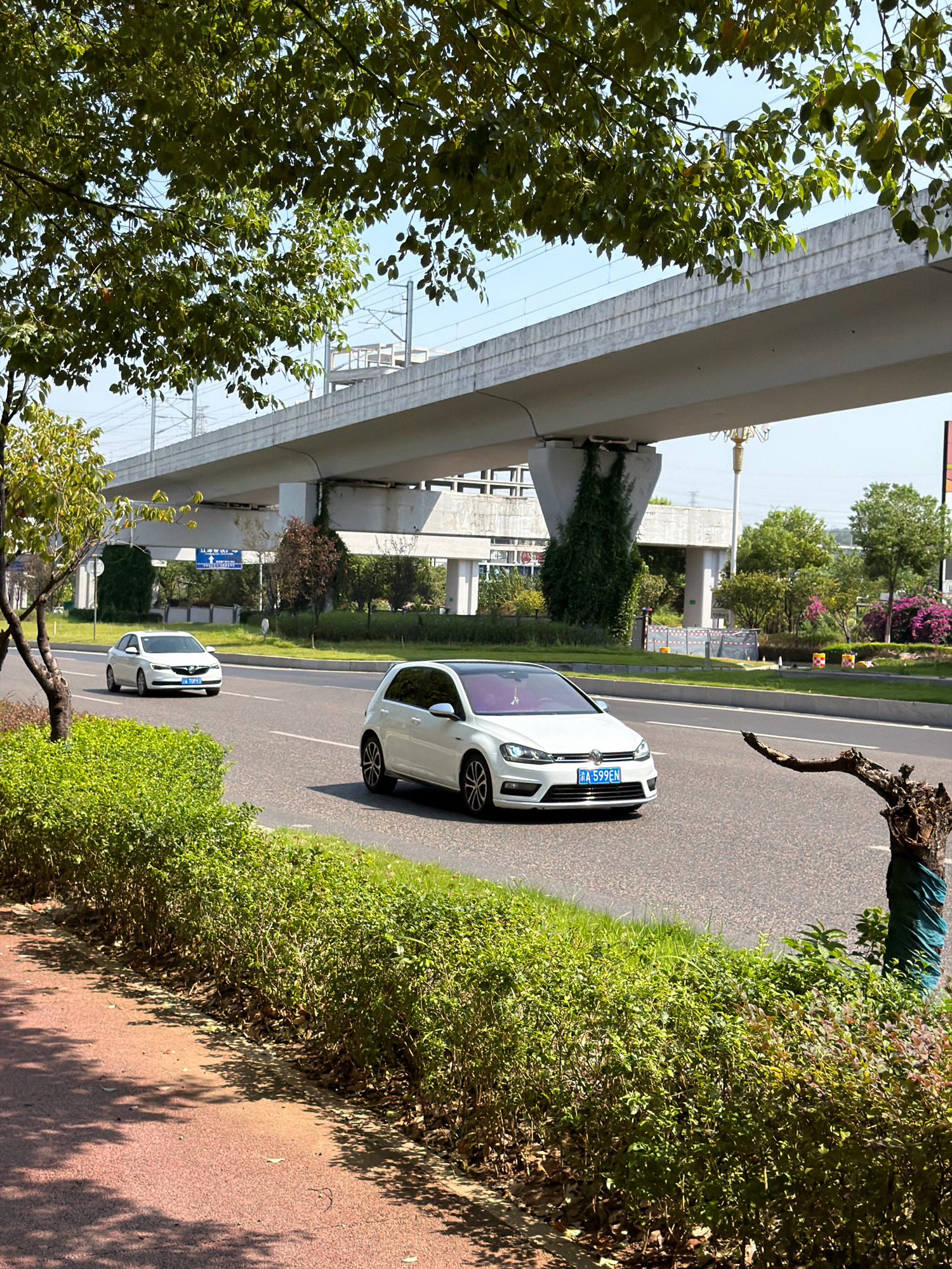 The image depicts a street scene in Jiangjin, China. A white Volkswagen Golf hatchback is driving on a paved road, with a white sedan in front of it. Both cars are traveling in the same direction. The road is flanked by manicured green hedges and a red-colored pedestrian walkway. Overhead, a large elevated structure, likely an overpass or a section of an elevated railway, casts shadows on the road and surrounding vegetation. The sky is clear and blue. The environment appears to be urban, with hints of buildings and infrastructure visible in the background, along with trees and flowering bushes. The overall impression is a sunny day in a well-maintained urban area.