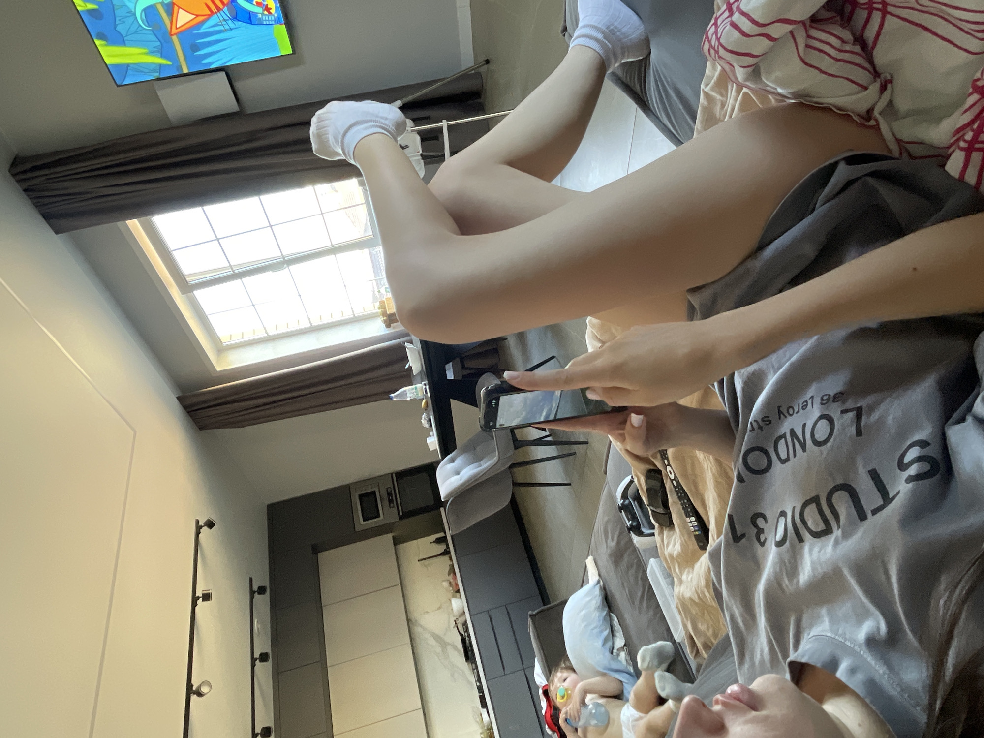A person wearing white socks is lying on a bed in a room with a window and a television. The person is holding a cell phone in their hands and taking a picture. A child is lying on the bed near the person. The room is in an apartment building with a kitchen. The city is Эстачи, Russia.
