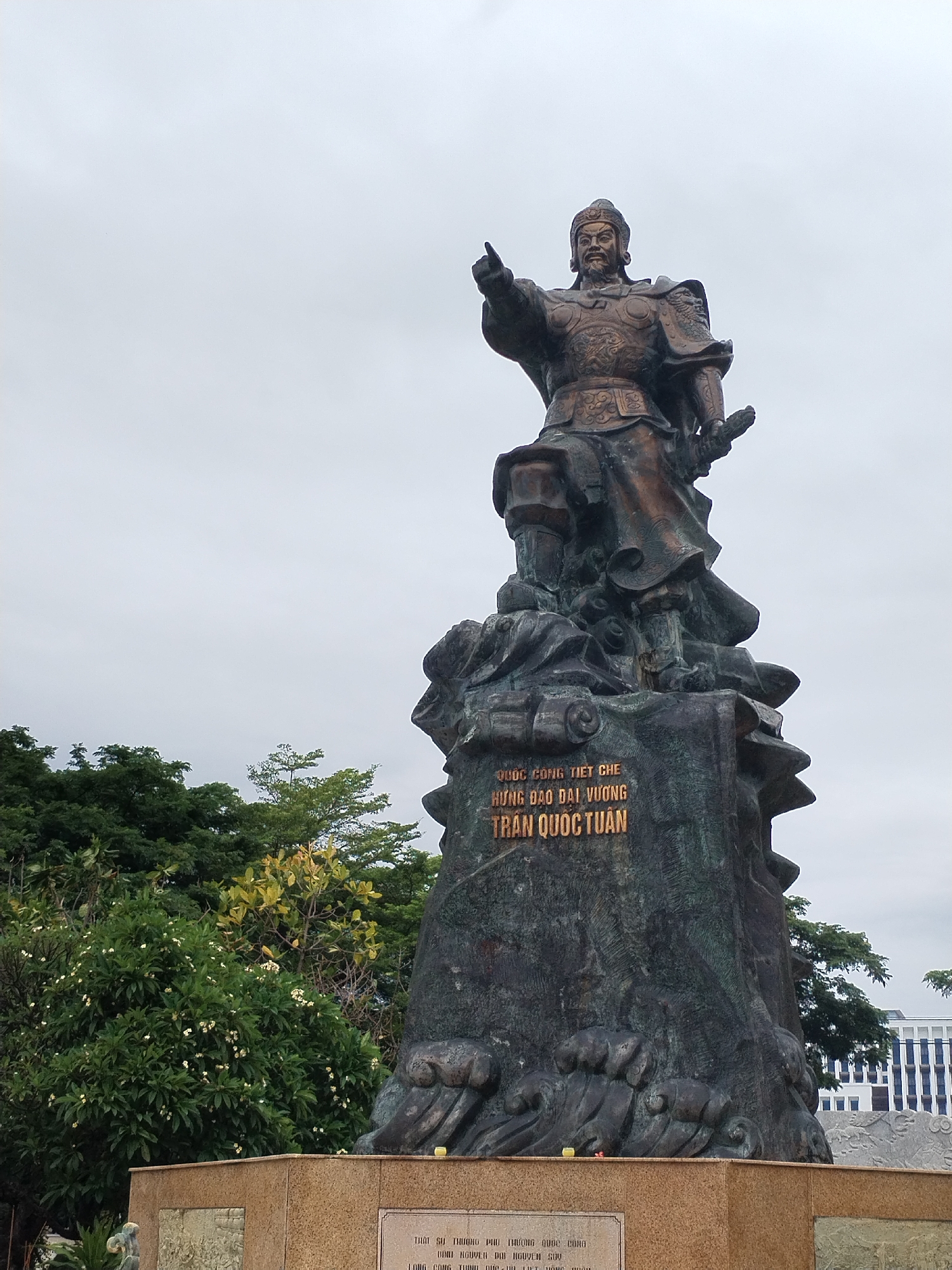 The image shows a bronze statue of Trần Quốc Tuấn, a Vietnamese general, situated in Nha Trang Ward, Vietnam. Trần Quốc Tuấn is depicted standing on a large stone pedestal, pointing his right hand forward. He is dressed in what appears to be traditional military garb. The base of the statue has an inscription in Vietnamese. The statue stands in front of a variety of green trees and a light gray sky. A granite block with inscriptions and bas-relief carvings makes up the lower portion of the base. In the background, there's a building with a white and blue facade.