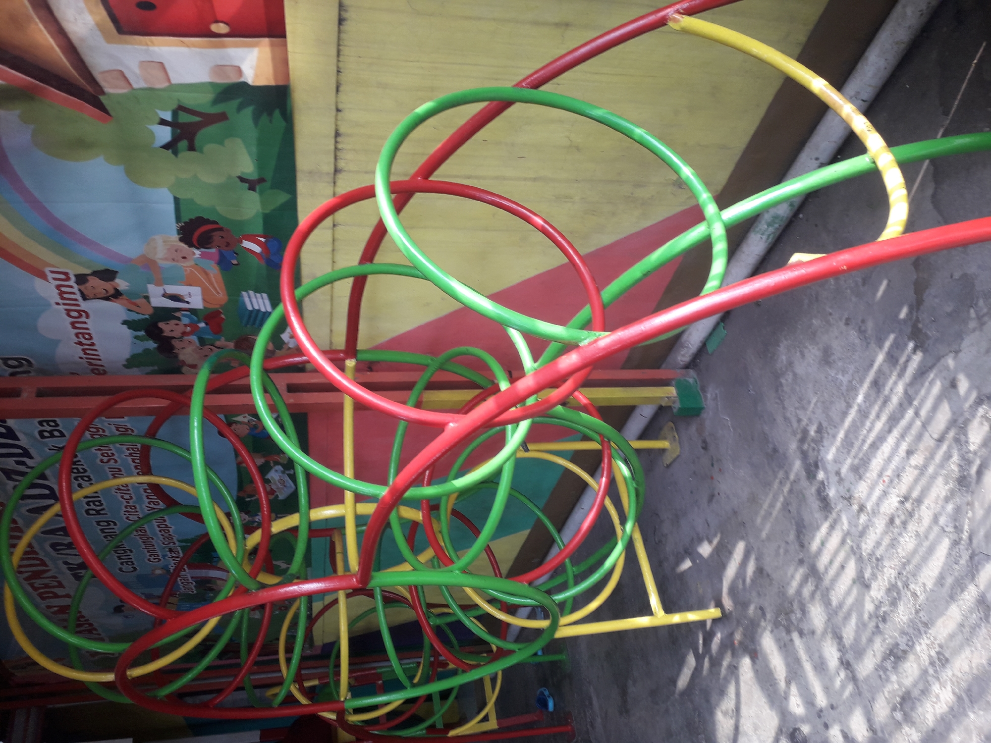 The image shows a colorful playground structure made of red, green, and yellow tubes, likely for children to climb and play on. The playground is situated outdoors on a concrete surface with shadows cast across it. In the background, there is a mural painted on a wall depicting children and a teacher reading a book, with a rainbow arching overhead and a drawing of a house. The mural appears to be part of a kindergarten or early childhood education facility, possibly located in Cangkang, Indonesia, as suggested by text on the mural itself which mentions "Cangkang RAUDZ-DZ" and "Cangkang, Indonesia". The overall scene is bright and cheerful, indicative of a place designed for children's activities.