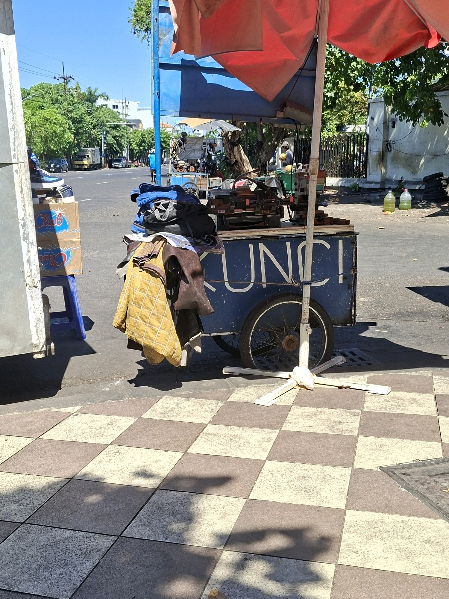 A cluttered street cart with the partial word "KUNCI" visible on its side is parked on a sidewalk. The cart is loaded with various items including clothing, tools, and what appear to be boxes. A red umbrella is open above the cart. In the background, a street with traffic and trees is visible, suggesting an urban setting. The sidewalk is paved with checkered tiles. The scene appears to be set in the city of Surabaya, Indonesia, based on visual cues.