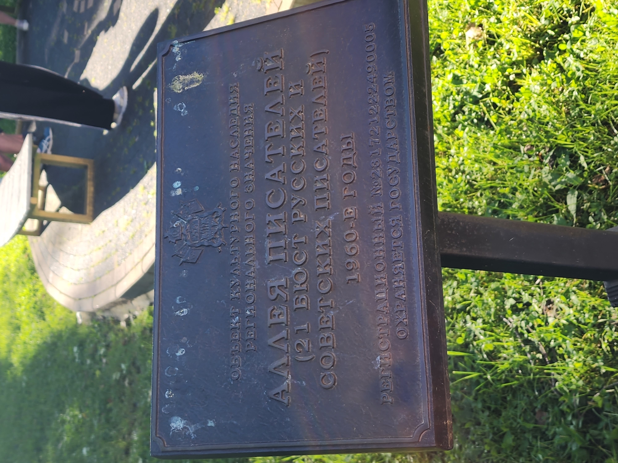 The media file captures an outdoor scene, primarily focusing on a dark, rectangular metal plaque affixed to a stand, set against a backdrop of lush green grass and paving. The plaque, likely made of bronze or a similar material, features raised Cyrillic text and a small embossed emblem at the top left. Scattered light green/white spots, possibly from oxidation or environmental exposure, are visible on its surface.
The scene depicts a sunny day, evidenced by prominent shadows cast on the ground, including long shadows of people and perhaps trees. In the upper left, partial views of a person's legs and feet are visible, along with a glimpse of a light-colored bench or table with a golden-colored frame. The ground beneath the plaque appears to be a mix of paved area and natural green grass. The overall setting suggests a public park, alley, or memorial area.
The visible text on the plaque, translated from Russian, reads:
"CULTURAL HERITAGE OBJECT
OF REGIONAL SIGNIFICANCE
ALLEY OF WRITERS
(21 BUSTS OF RUSSIAN AND
SOVIET WRITERS)
1960S
REGISTRATION NO. 231721222490005
PROTECTED BY THE STATE"
This plaque indicates that the location is an "Alley of Writers" established in the 1960s, featuring 21 busts of Russian and Soviet writers, and is designated as a cultural heritage site of regional significance, protected by the state in Sochi, Russia.