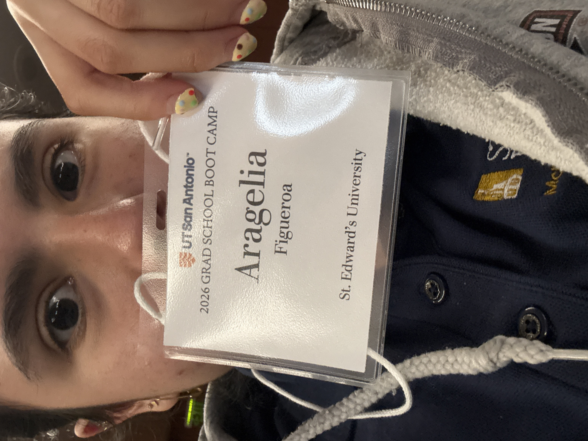 A person is holding up a conference badge, likely at an event in San Antonio, Texas. The badge identifies the individual as "Aragelia Figueroa" and states they are associated with "St. Edward's University." The badge also indicates attendance at the "2026 GRAD SCHOOL BOOT CAMP" hosted by "UT San Antonio." The person is wearing a dark blue collared shirt, partially obscured by a gray hooded sweatshirt.
