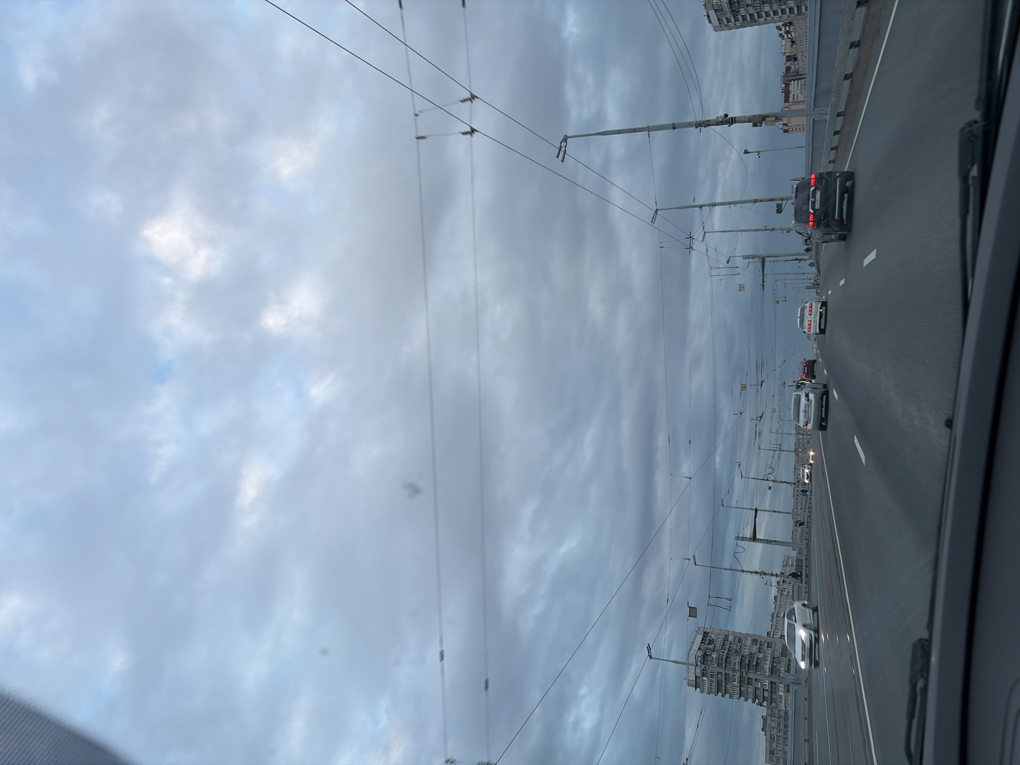 The image shows a view from inside a car driving on a multi-lane road. The sky is overcast with gray and white clouds. On the right side of the road are buildings and lampposts. There are several cars on the road. The location is Saint Petersburg, Russia.