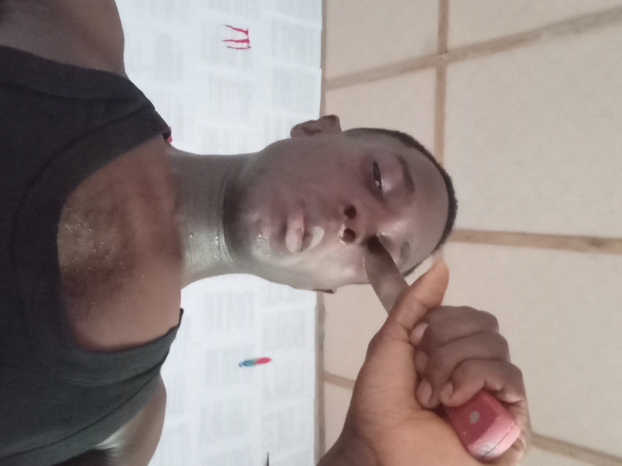 A close-up, low-angle shot of a man with dark skin wearing a dark tank top. The man is holding a small, pink object near his face. His skin appears shiny, possibly sweaty or wet. His expression is somewhat serious. The background includes a wall with a tiled or paneled appearance. Red and blue marks are visible on a white surface in the background. The location is claimed to be Akwete, Nigeria, but there is no corroborating evidence in the image itself to confirm this.