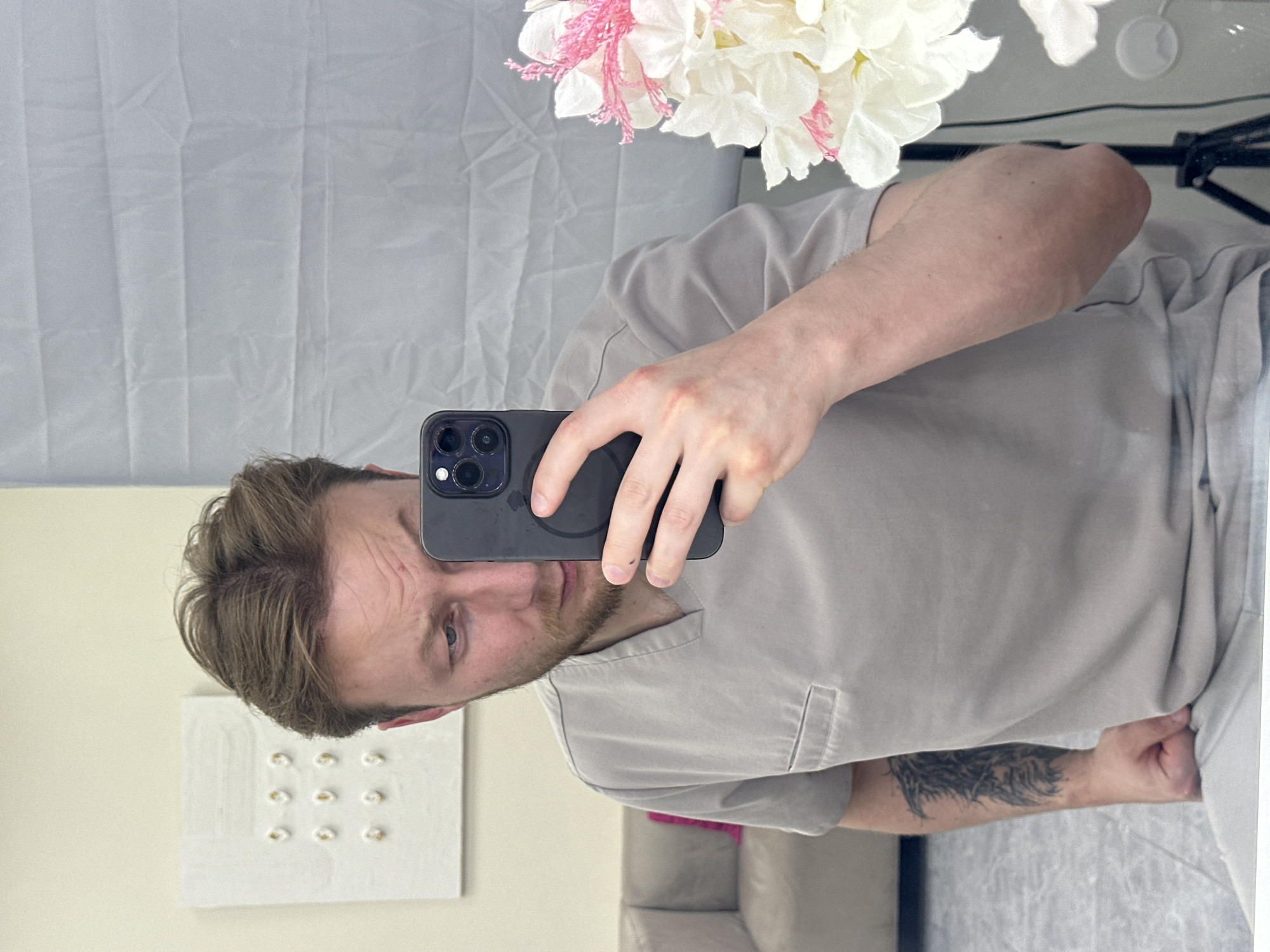 An indoor mirror selfie captures a light-skinned male with blonde-to-light-brown hair and a short beard, wearing a light gray/beige short-sleeved top with a v-neck and a chest pocket. The image is presented rotated 90 degrees clockwise, so the man appears lying on his side. He holds a dark-cased smartphone, likely an iPhone due to its distinctive triple camera array, in front of his face, taking the photo. His gaze is directed towards the camera with a neutral expression, and slight wrinkles are visible on his forehead.
A prominent, intricate black tattoo with an abstract, feathery or wing-like design covers a significant portion of his left forearm.
The background visible in the mirror reflection includes several elements. At the top right (when the image is viewed upright), a large cluster of artificial white flowers with delicate pink accents is visible. Behind the man, a light grey, somewhat wrinkled fabric backdrop or sheet hangs. To his left, a small, white rectangular art piece featuring a grid of nine golden, circular, textured elements is mounted on a light cream-colored wall. A light-colored sofa or upholstered furniture is partially reflected in the lower right background. Some wires and part of a black stand are also faintly visible. There is no visible text in the image. The setting is an indoor space, potentially a home or studio, located in Sochi, Russia.