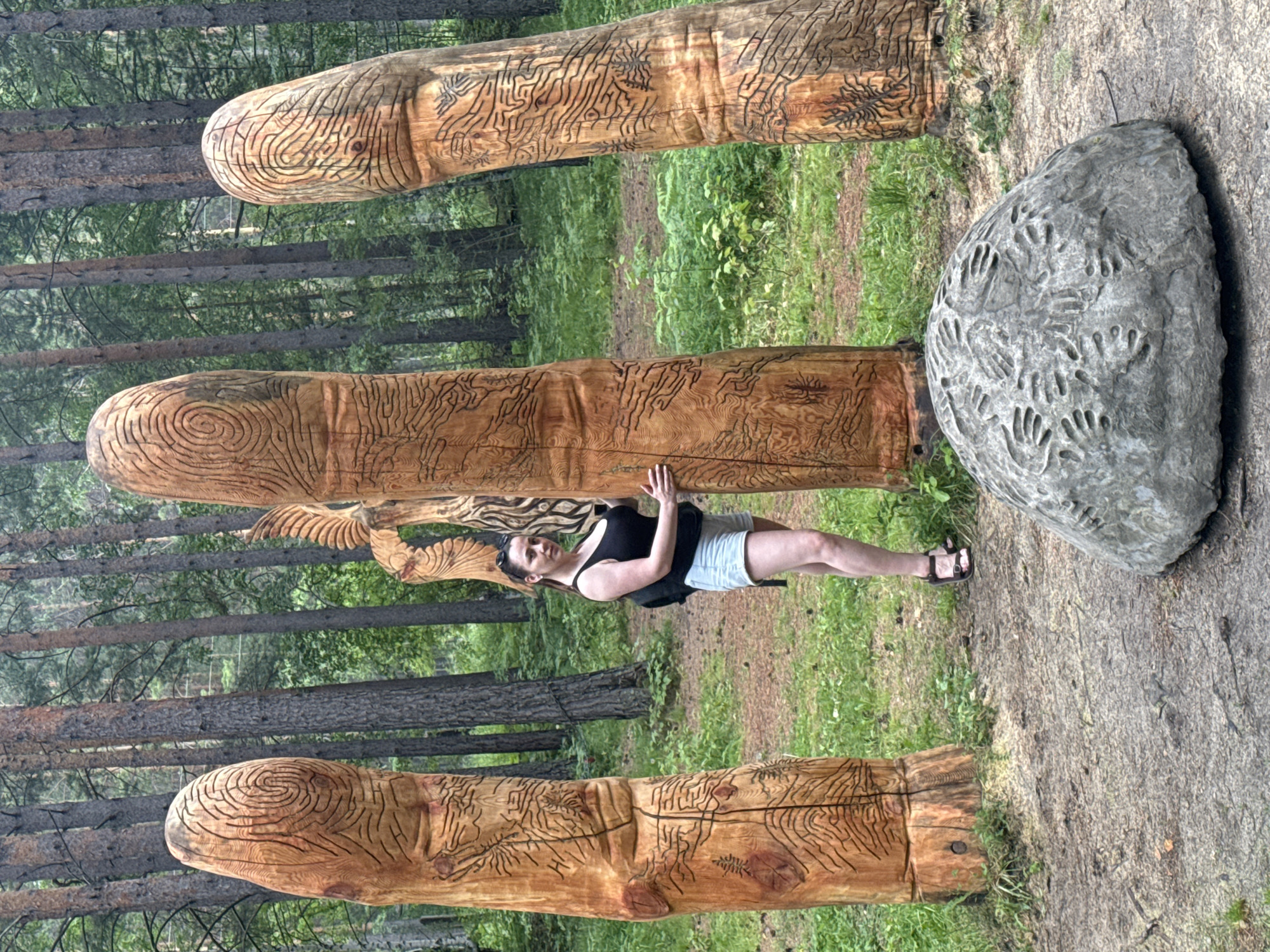 The image depicts an outdoor scene in a forest, possibly near the city of Savvateyevka, Russia. In the foreground, a large, rounded gray stone is visible, imprinted with numerous handprints. Behind the stone, three tall, wooden sculptures resembling fingers stand upright. These sculptures are intricately carved with patterns, including what appears to be a fingerprint design on the top of each. A woman, wearing a black tank top and light-colored shorts, stands between two of the wooden sculptures. She is holding onto the sculpture on the right, facing the camera. The background is dominated by a dense forest of tall pine trees and green foliage.