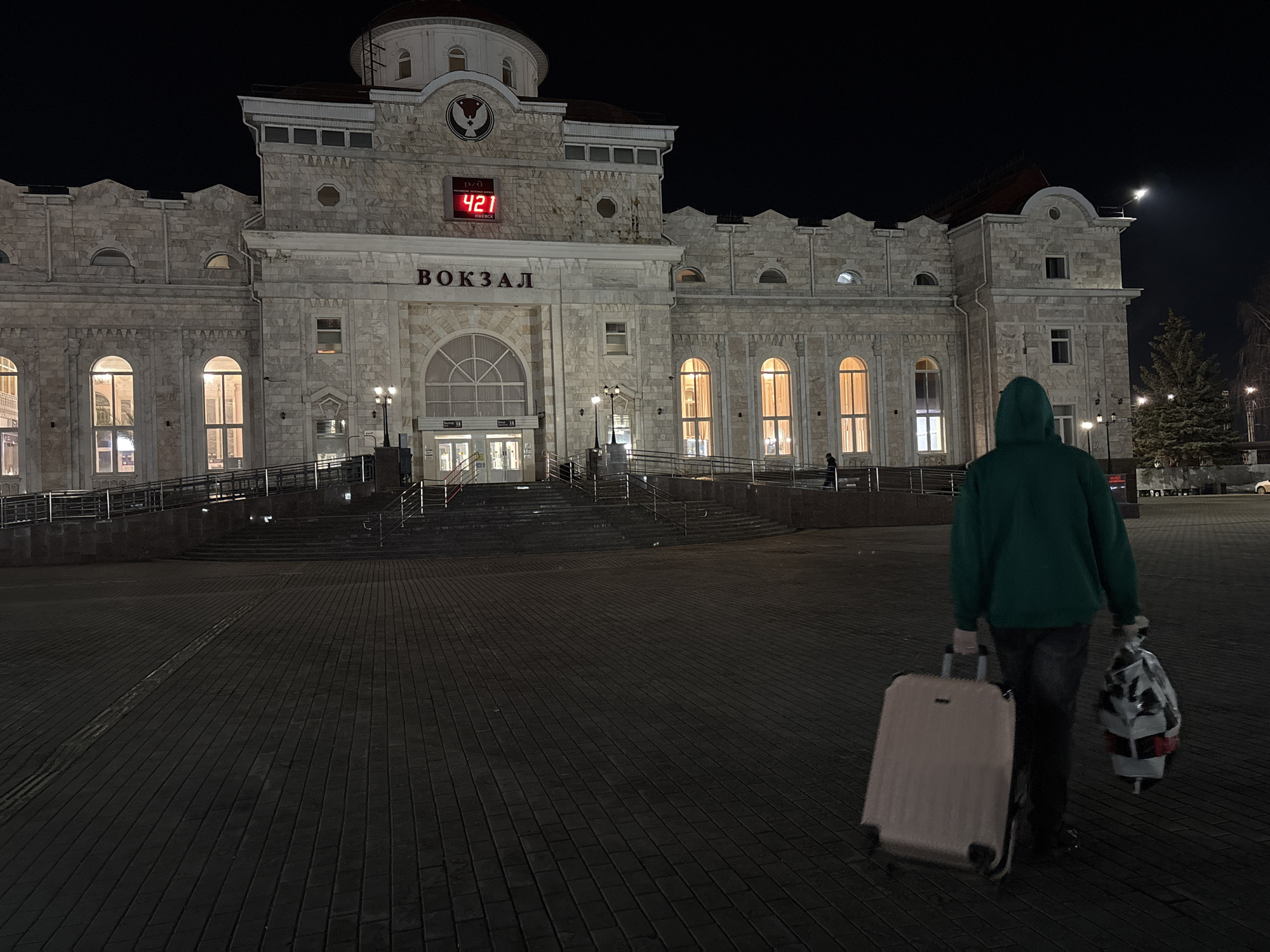 At night, in Izhevsk, Russia, a person in a green hooded sweatshirt walks away from the train station, Вокзал (Vokzal). The individual is pulling a light-colored suitcase and carrying a white plastic bag with black and red accents. The station is lit by interior and exterior lights, showing a large brick structure with arched windows. A digital clock above the station's entrance displays the time as 4:21. The moon is visible in the upper right sky. The ground is brick paved.