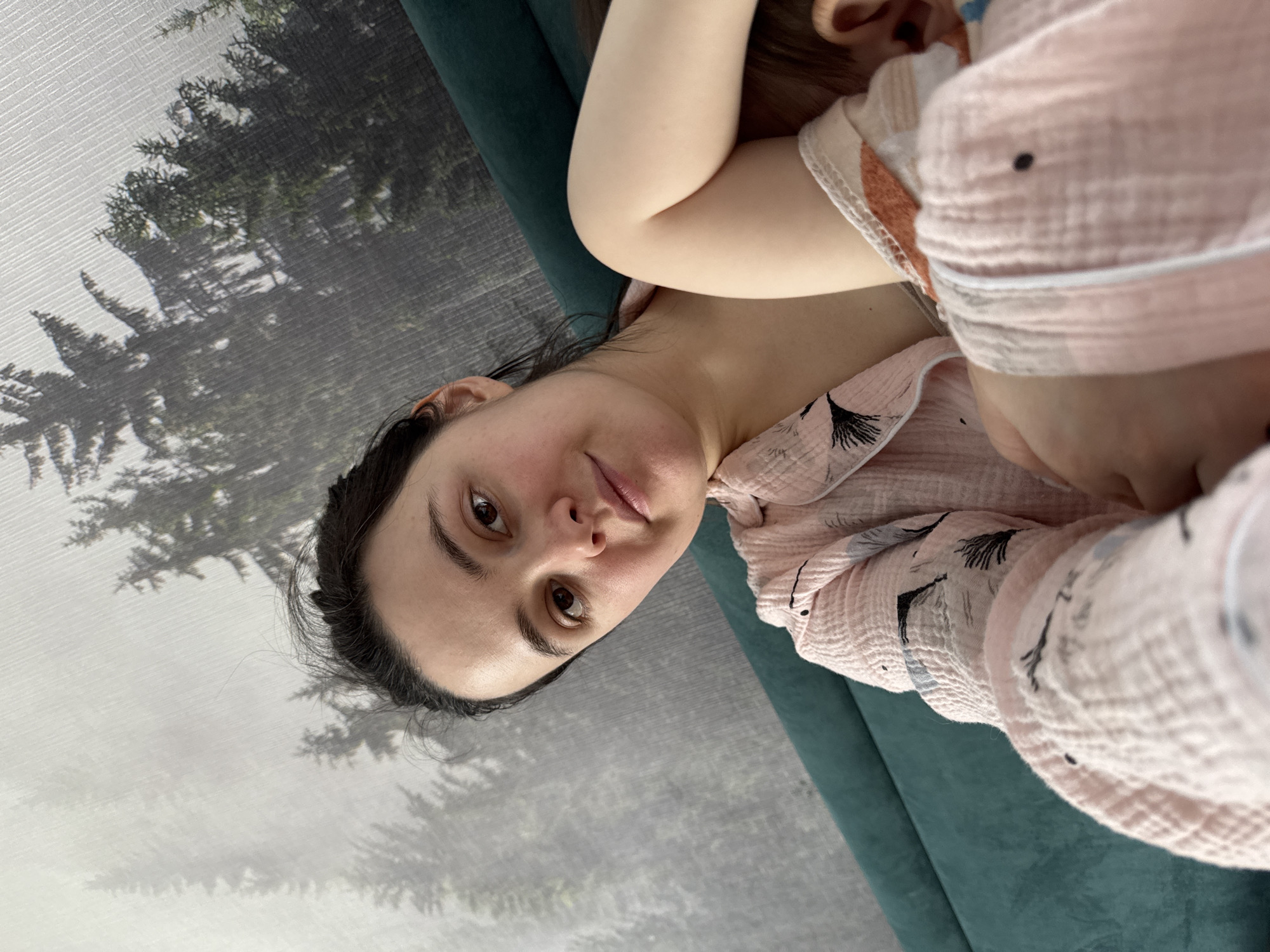 A woman is lying on a green couch in front of a gray wall. The wall has a picture of trees on it. The woman has dark hair and brown eyes. She is wearing a pink shirt. A baby is lying on her chest. The location cannot be determined based on the information provided.
