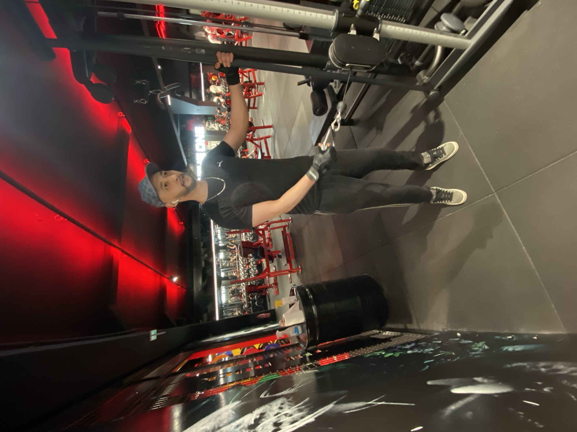 A man wearing a black shirt, black pants, grey gloves, and grey sneakers is photographed in a gymnasium. He also wears a blue cap and a silver necklace. He is holding onto a cable machine with his left hand and appears to be pulling on the cable with his right hand, presumably working out. The gym features red-accented walls, various exercise machines, and a grey floor. A black trash can is situated near the base of the image. The location is likely in Strasbourg, France, given the assumption.