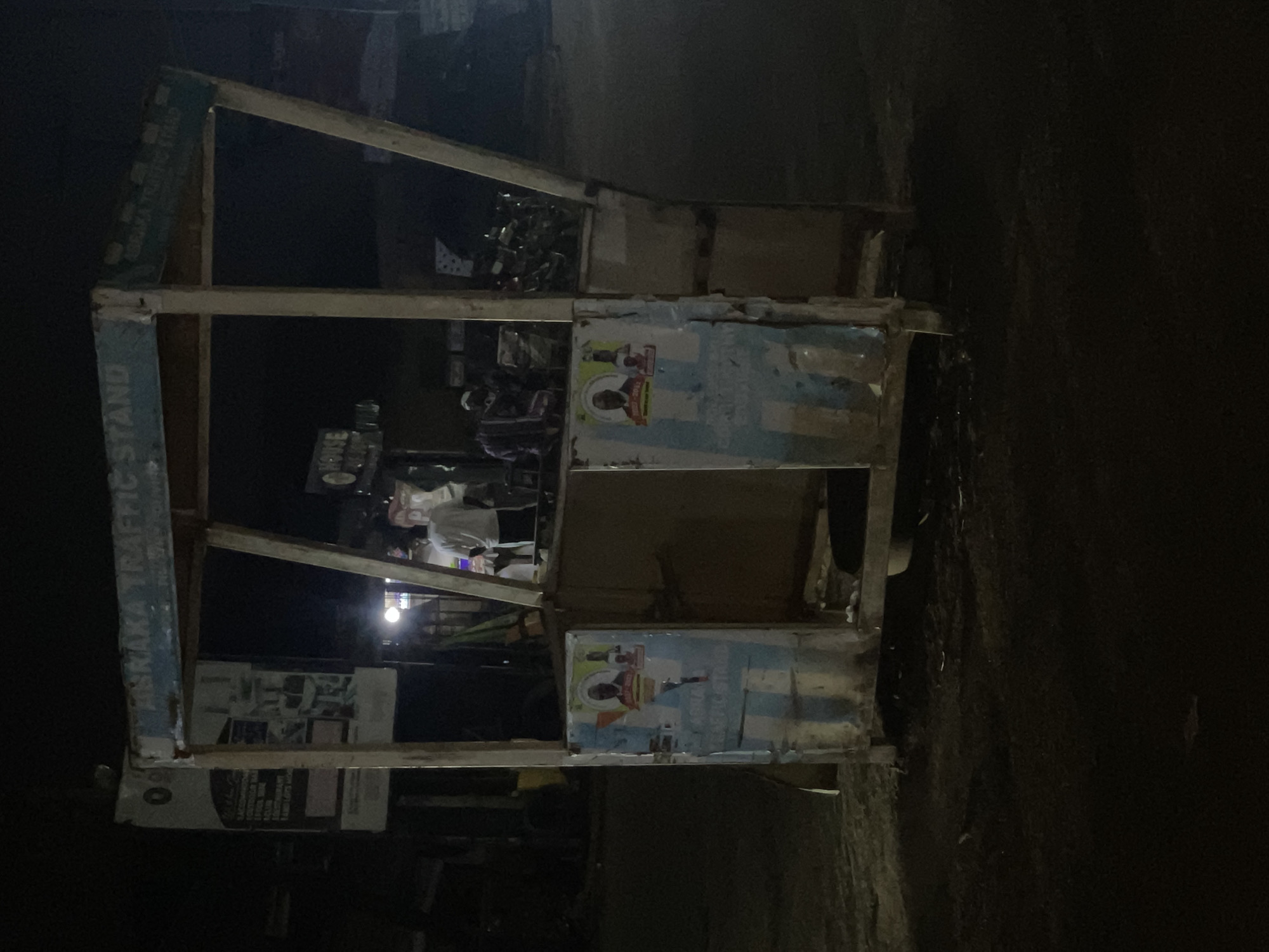 A poorly lit, upright-oriented image shows a weathered metal and wood vendor stand at night in Abraka, Nigeria. The words "ABRAKA TRAFFIC STAND" are partially visible on the left vertical support. The stand is multi-tiered, with various items inside including what appears to be packaged goods, personal items, and possibly food items. The front panels of the stand are partly covered with faded posters, one showing a man's portrait. In the background, a figure is partially visible, seemingly a person attending to the stand. The area around the stand appears dark and nondescript.