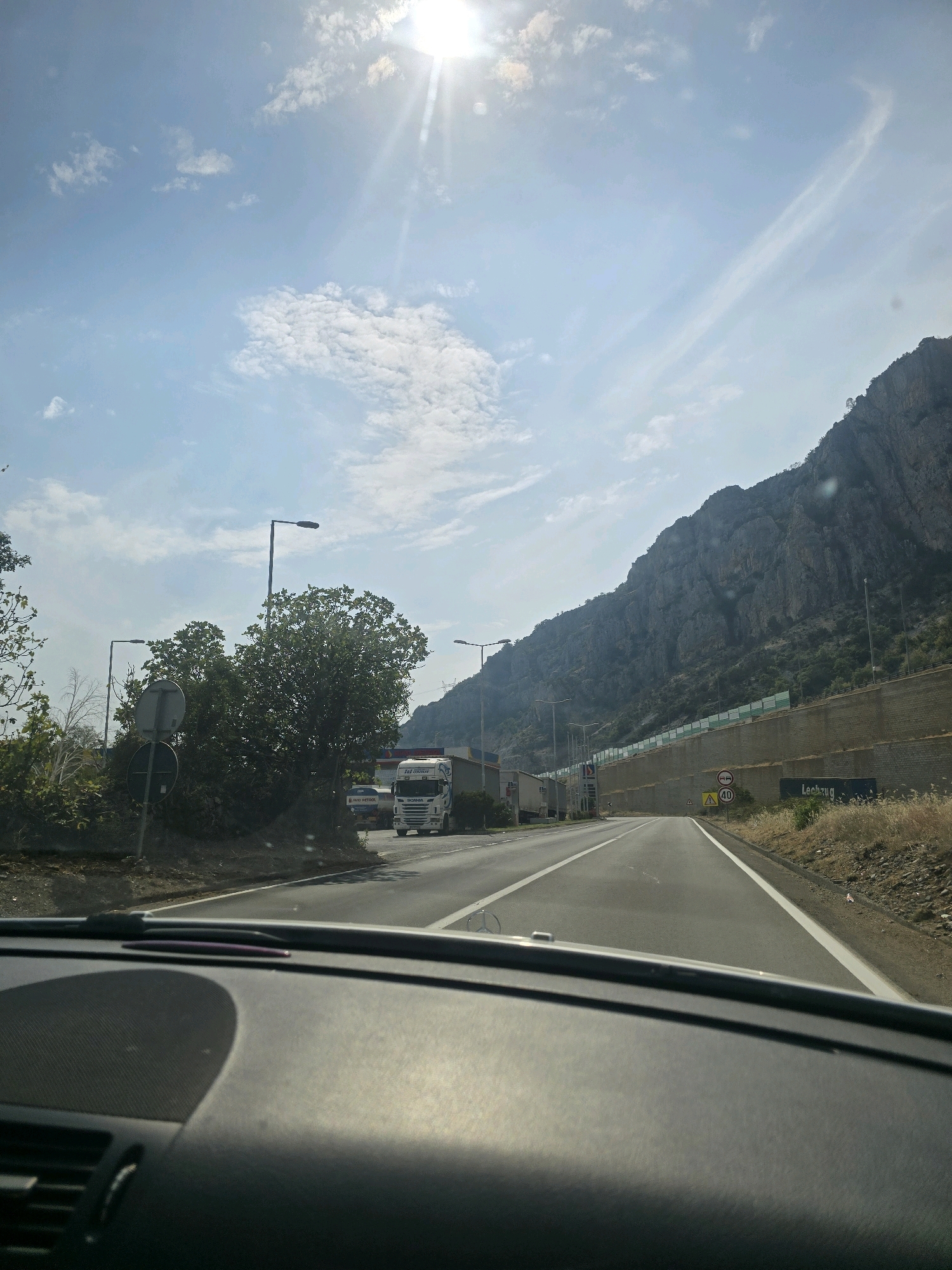 The image, taken from the interior of a vehicle, depicts a road lined with a steep, rocky mountainside on the right and vegetation and infrastructure on the left. The road itself is paved, with lane markings visible. Several trucks are present on the road, some appearing to be stopped or moving slowly. Signs are visible, including a speed limit sign indicating 40, and what appears to be a "no entry" sign. A large retaining wall separates the road from the mountainside. The sky is blue with scattered white clouds and the sun is shining brightly, suggesting daytime. The location is identified as being on the road to Podgorica, Montenegro.