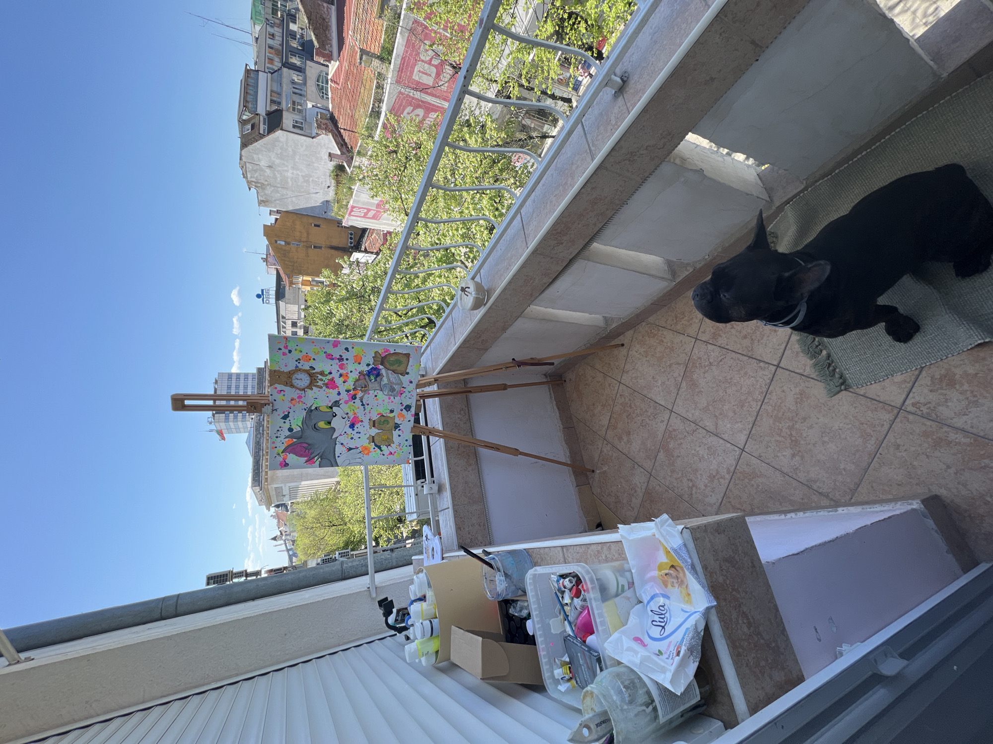 An eye-level photograph, rotated clockwise, depicts an outdoor balcony painting setup in Burgas, Bulgaria, as deduced from the buildings visible in the background. A black French bulldog is lying on a small green rug in the top right corner. An easel holds a colorful painting with Tom and Jerry characters. Various art supplies including paint tubes, containers, and wipes are scattered on a table to the right of the easel. A white railing separates the balcony from the cityscape. The sky is clear and blue. Buildings of various heights are visible in the background.