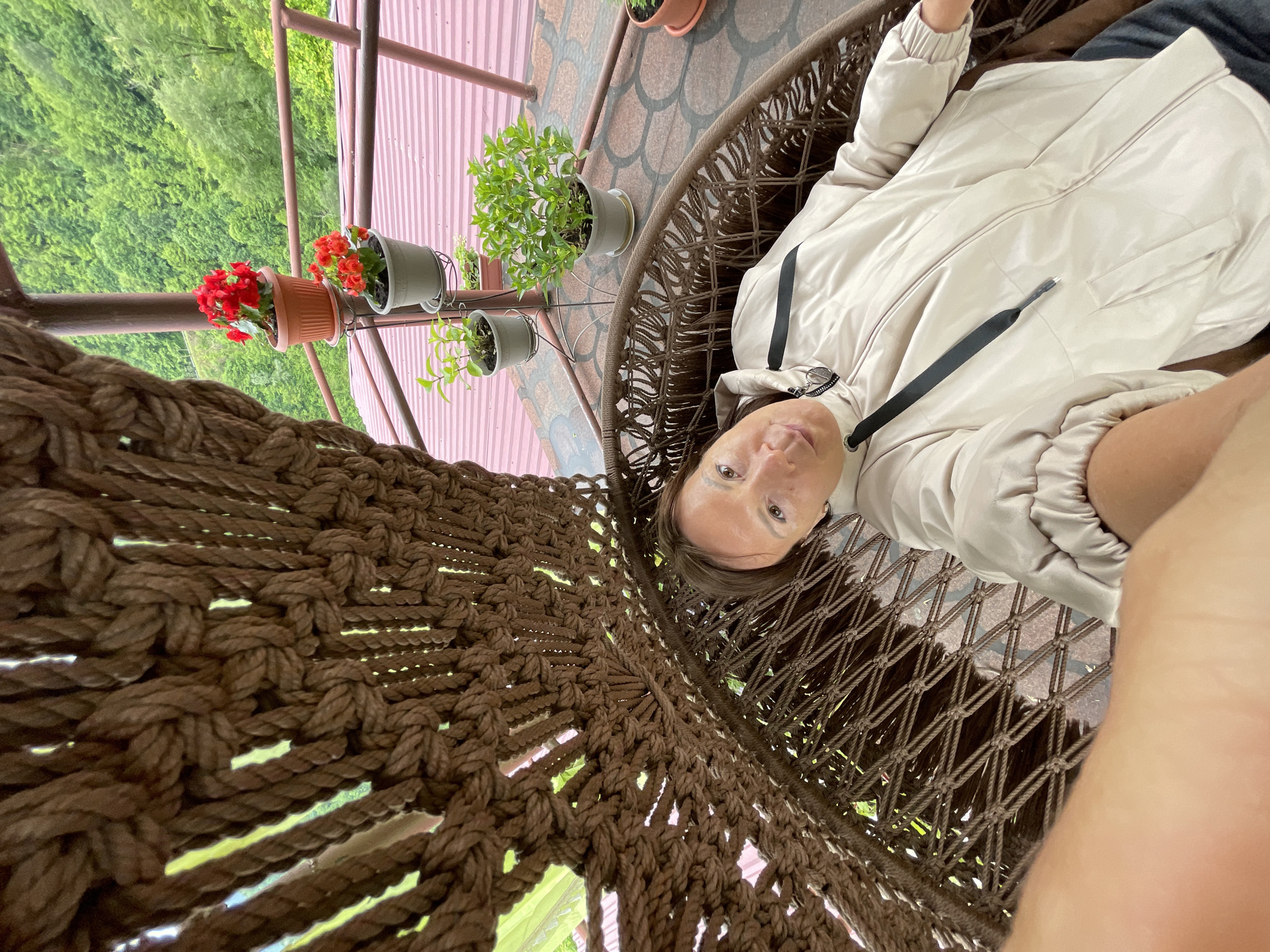 A medium, high-angle shot shows a person in a brown macrame hanging chair on a porch. The person, appearing to be a light-skinned woman, is reclining in the chair and looking up at the camera. She is wearing a light-colored jacket with black drawstrings. Overhead, there is a pink corrugated metal roof with a railing and several potted plants including red flowers. Green trees and a forest are visible through the macrame and beyond the roof. The location is presumed to be in or around Khartsyz Pervyi, Russia.